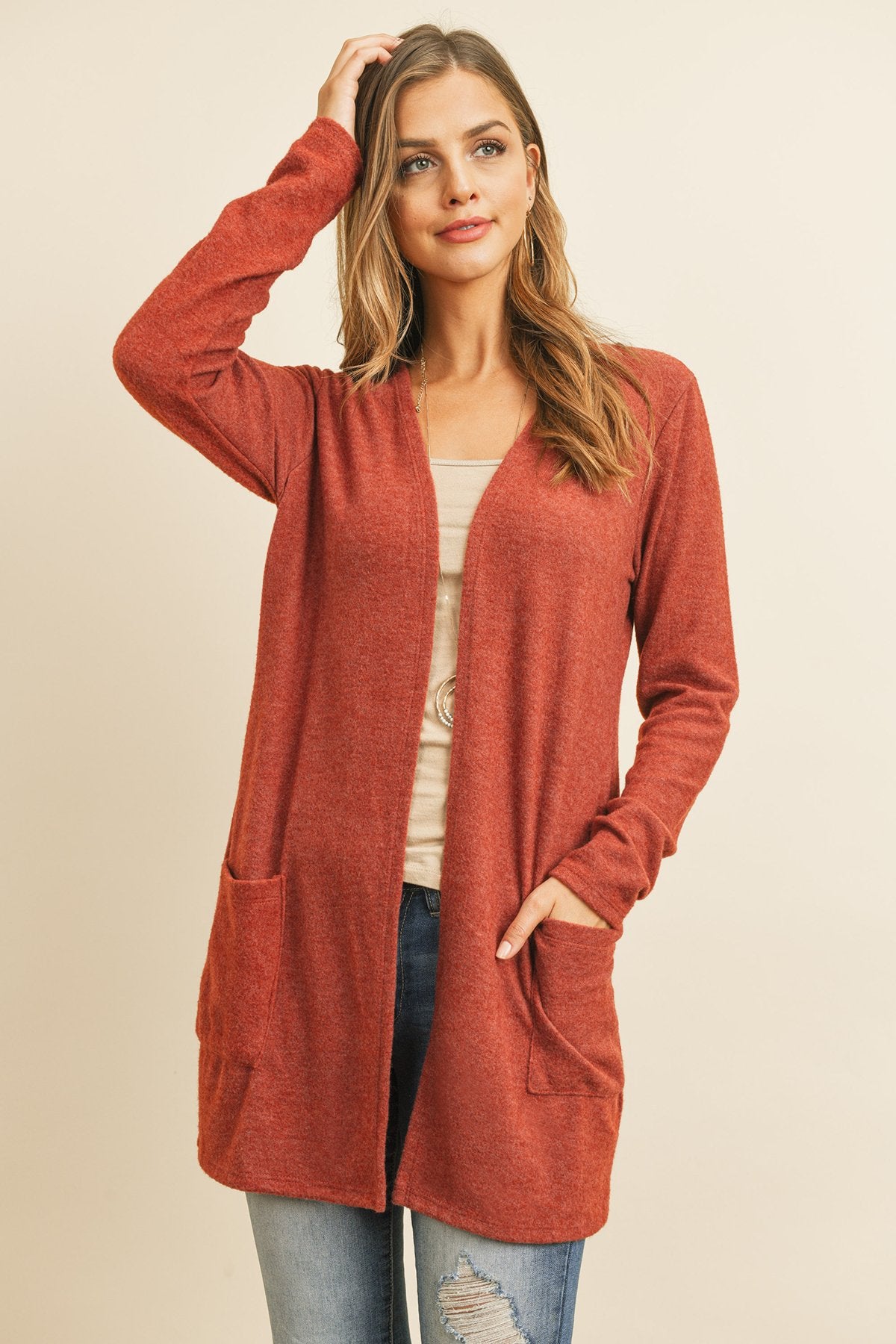 A woman in a teal Solid Long Sleeved Open Front Pocket Cardigan by Riah Fashion over a beige top stands against a pink background, smiling with hands in pockets.