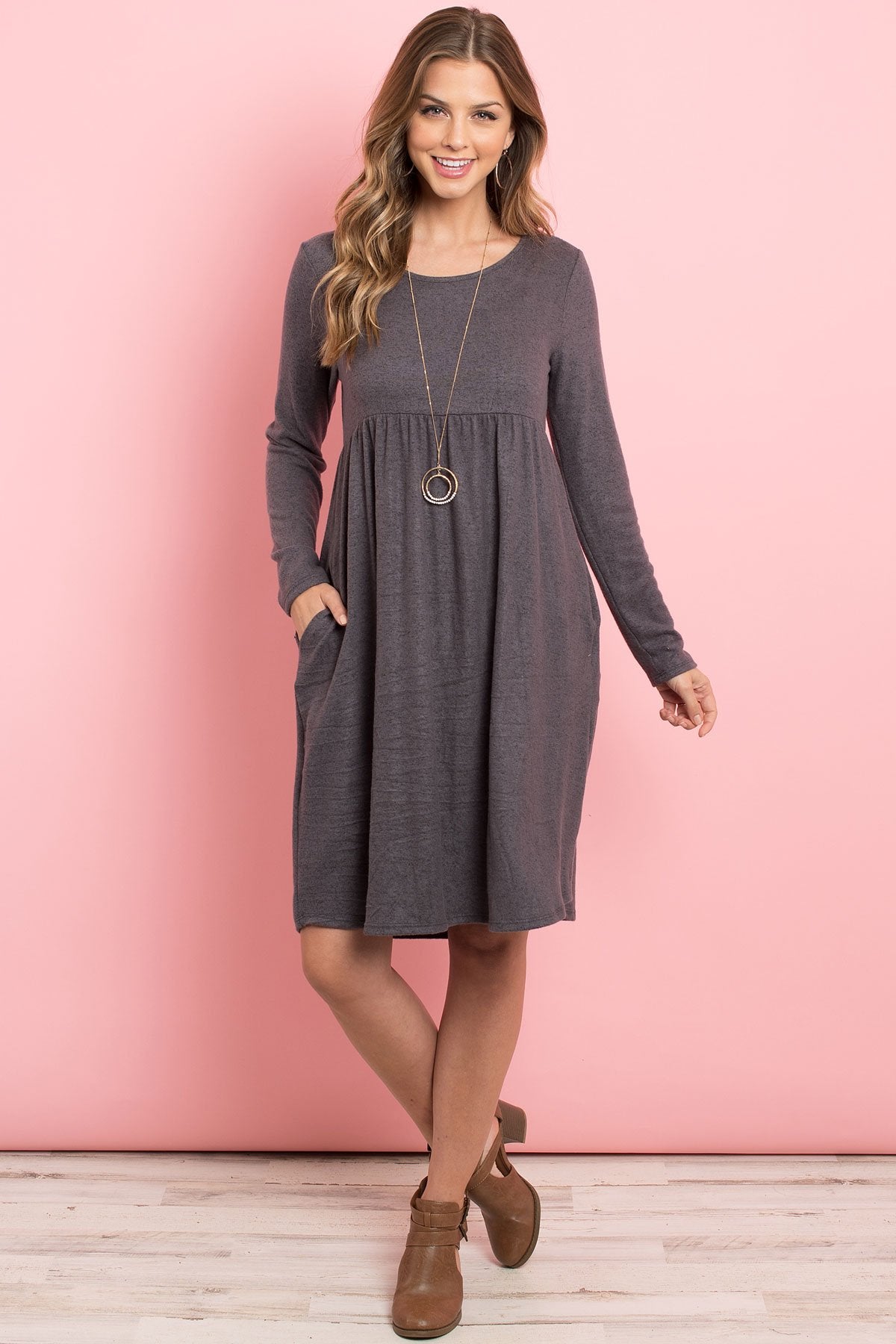 A woman in a Riah Fashion Boat Neck Brushed Hacci Round Hem Dress in Dark Ruby stands before a pink backdrop, adorned with a long necklace featuring circular pendants.