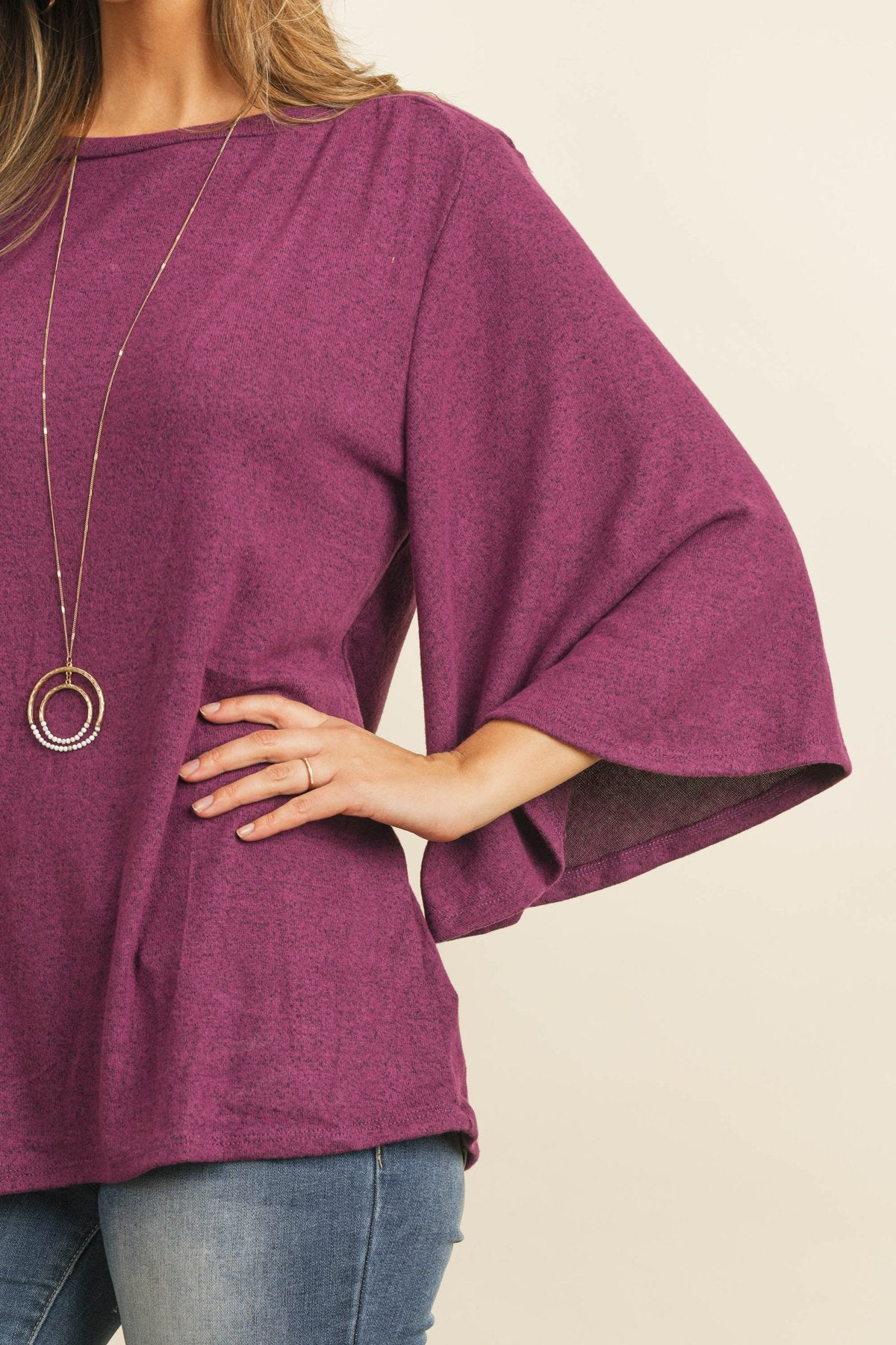 A person in a Riah Fashion Boat Neck Bell Sleeve Solid Hacci Brushed Top and ripped jeans stands against a neutral background, accessorized with hoop earrings and a long necklace.