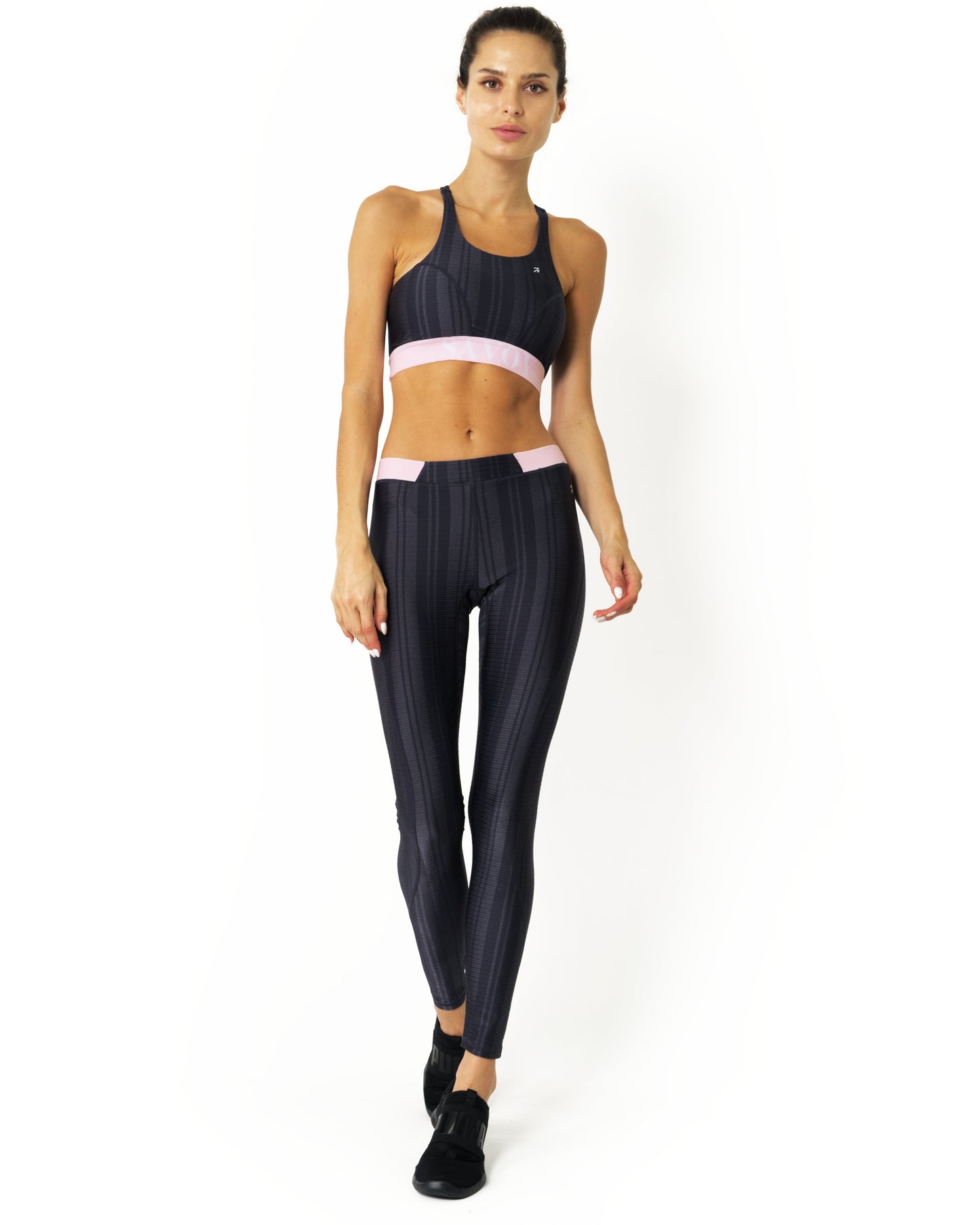 A person models the Savoy Active Greyson Leggings, featuring black and gray stripes with a pink waistband, paired with black shoes against a white background.