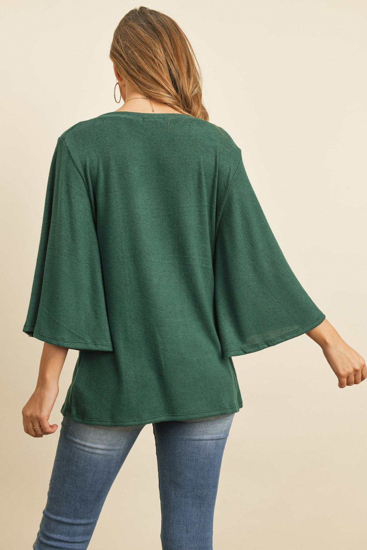 A person in a Riah Fashion Boat Neck Bell Sleeve Solid Hacci Brushed Top and ripped jeans stands against a neutral background, accessorized with hoop earrings and a long necklace.