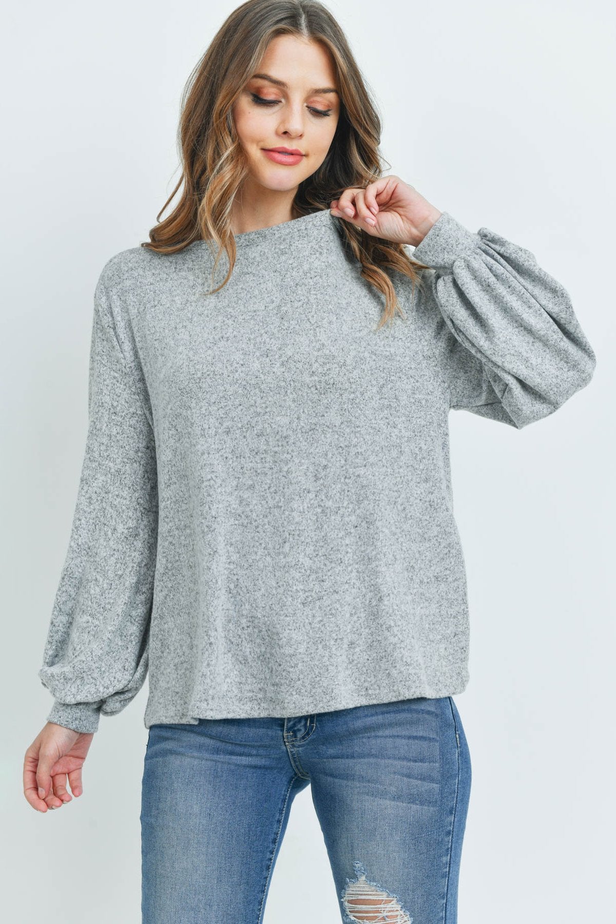 A woman models a Riah Fashion Puff Sleeved Boat Neck Two Toned Brushed Hacci Top with ripped jeans and brown boots on a concrete floor against a plain backdrop, accessorized with a long necklace.