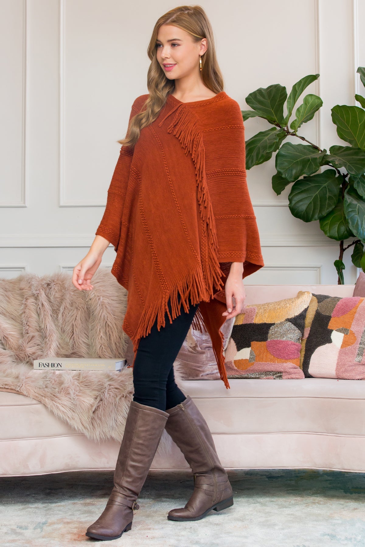 A person stands wearing a Riah Fashion Asymmetric Fringe Poncho in one color, paired with jeans and brown boots, smiling while holding the poncho. In the background are a plant and a sofa with cushions.