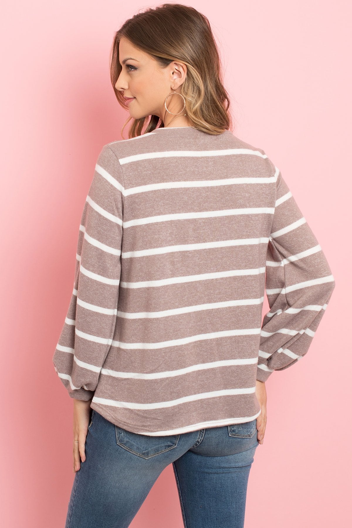 A woman in a Riah Fashion Striped Long Sleeved Round Neck Swing Top and jeans stands against a pink backdrop, wearing a long necklace and touching her hair.