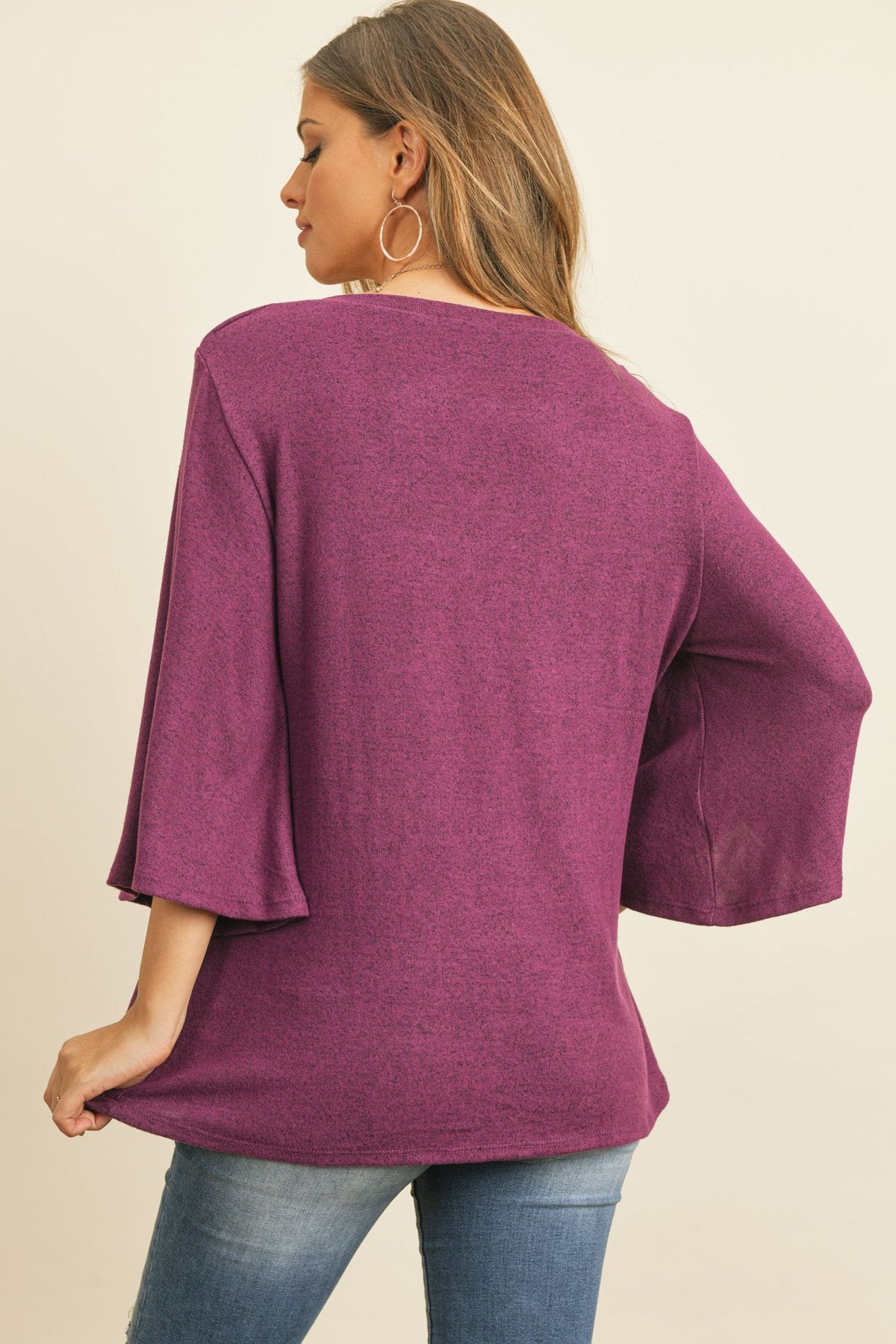 A person in a Riah Fashion Boat Neck Bell Sleeve Solid Hacci Brushed Top and ripped jeans stands against a neutral background, accessorized with hoop earrings and a long necklace.