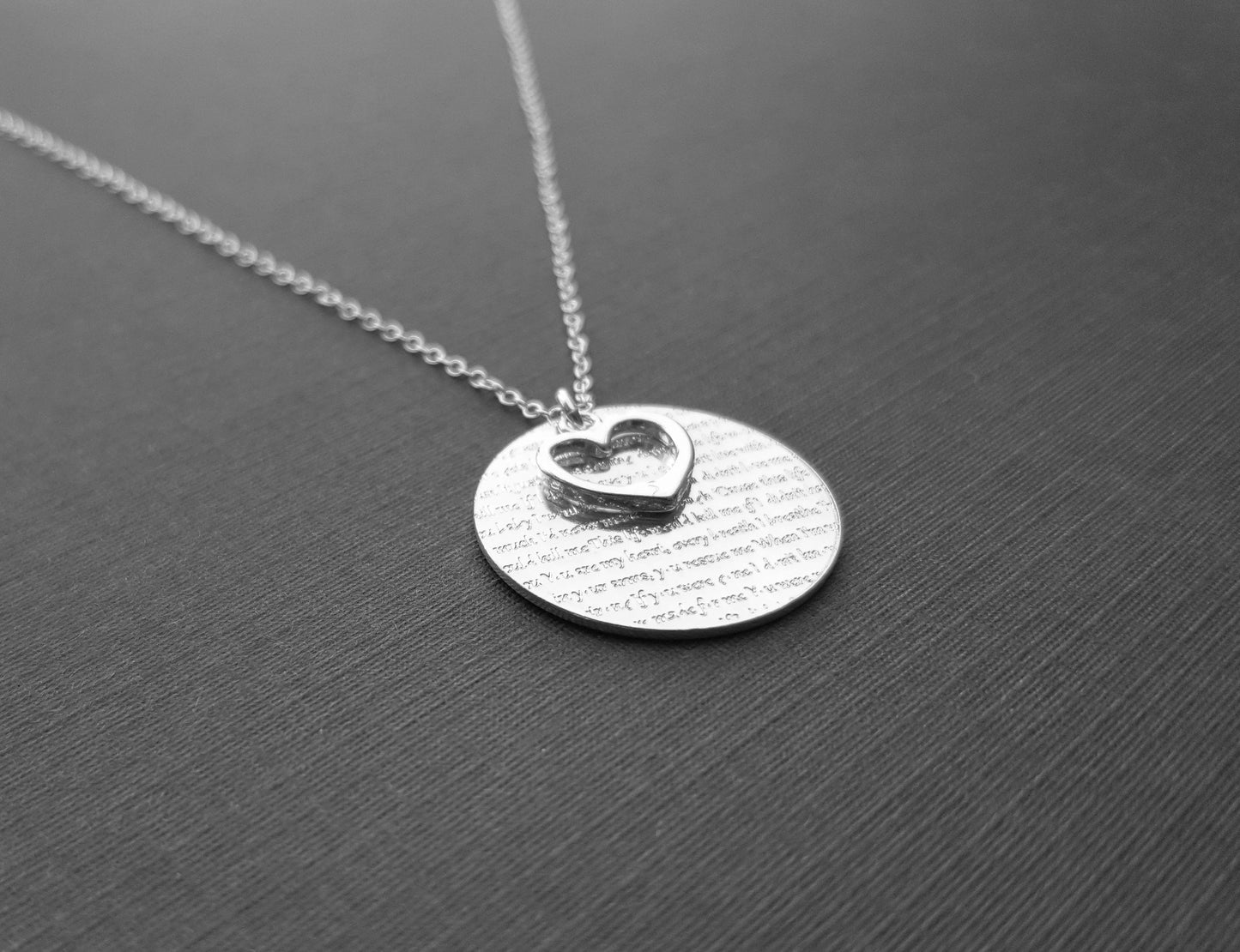 The Words of Love Necklace by Sincerely Silver is a silver piece with a round pendant showcasing printed text and an overlapping heart-shaped cutout.