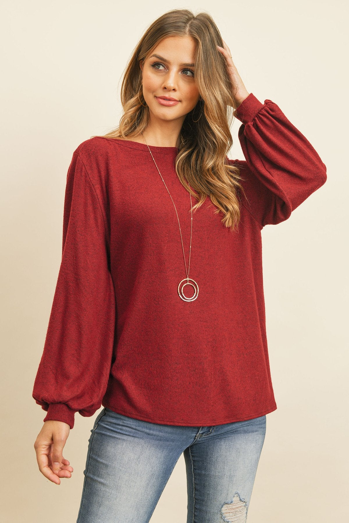 A woman models a Riah Fashion Puff Sleeved Boat Neck Two Toned Brushed Hacci Top with ripped jeans and brown boots on a concrete floor against a plain backdrop, accessorized with a long necklace.