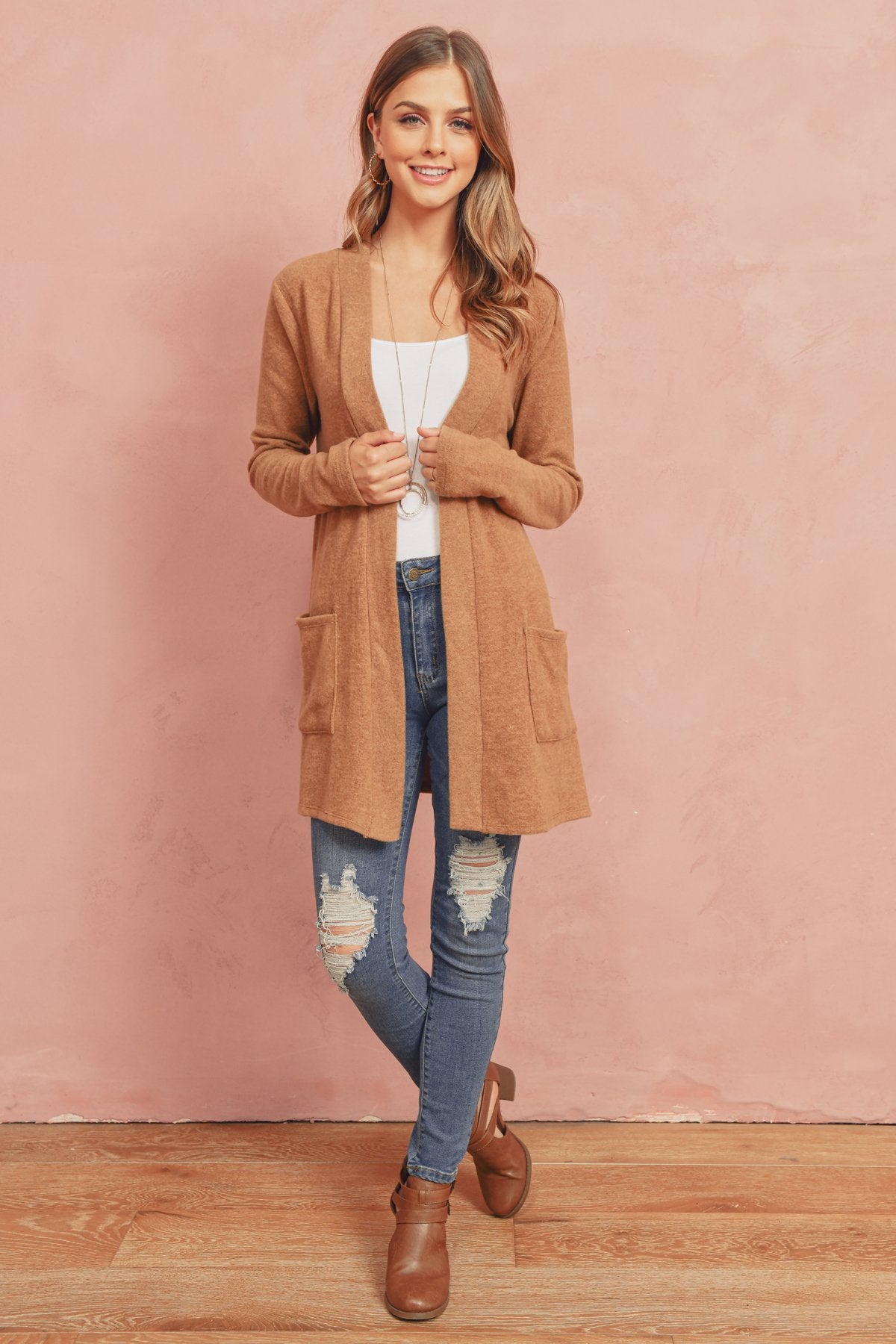 A woman in a Brushed Melange Open Front Cardigan by Riah Fashion and ripped jeans stands against a pink background, hands in her pockets.