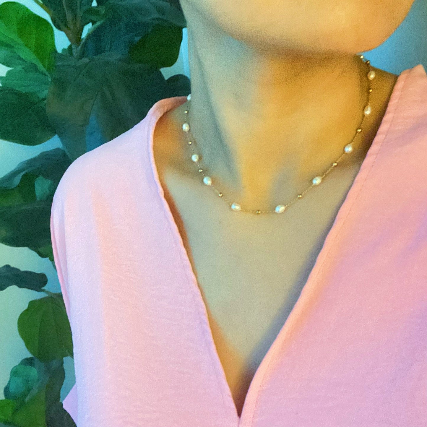 The Ashley Freshwater Pearl Necklace by Ellison + Young is a delicate gold chain adorned with small pearls and tiny gold beads, elegantly displayed on a light marble surface with green leaves and white stones.