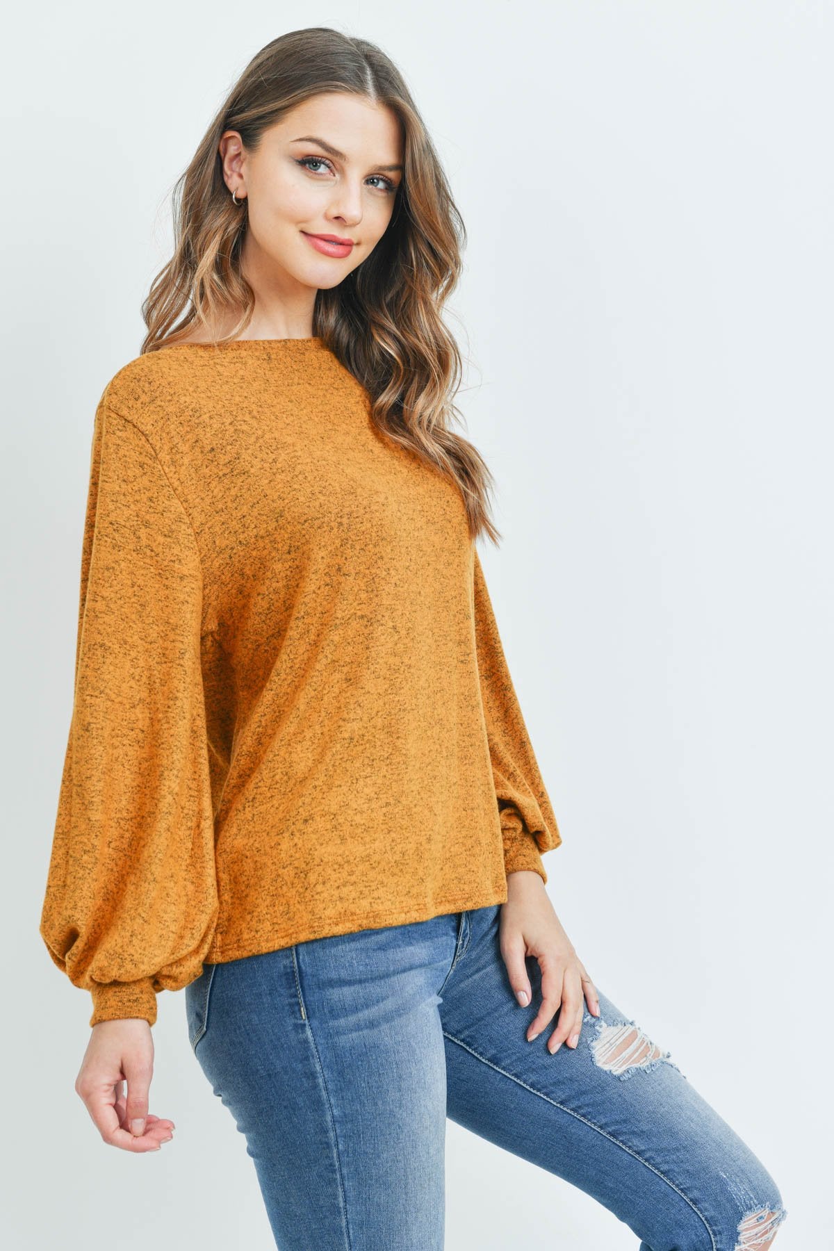 A woman models a Riah Fashion Puff Sleeved Boat Neck Two Toned Brushed Hacci Top with ripped jeans and brown boots on a concrete floor against a plain backdrop, accessorized with a long necklace.