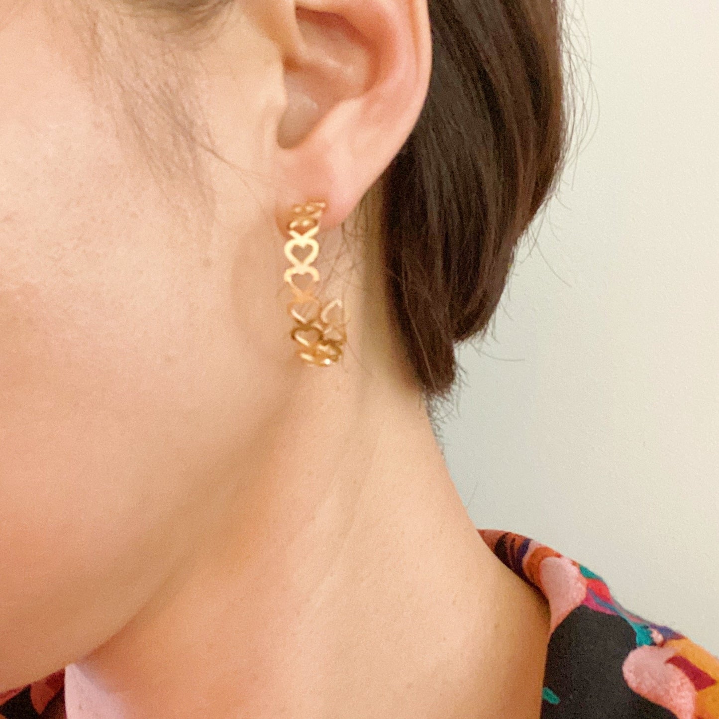 Ellison + Youngs Linked Heart Hoop Earrings, featuring heart-shaped cutouts, are elegantly displayed on a white marble surface surrounded by white pebbles.