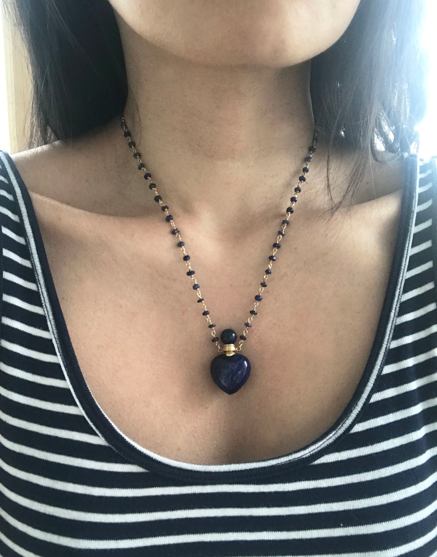 The Gena Myint Lapis Lazuli Heart Necklace features a blue beaded design with a heart-shaped pendant and gold chain clasp.