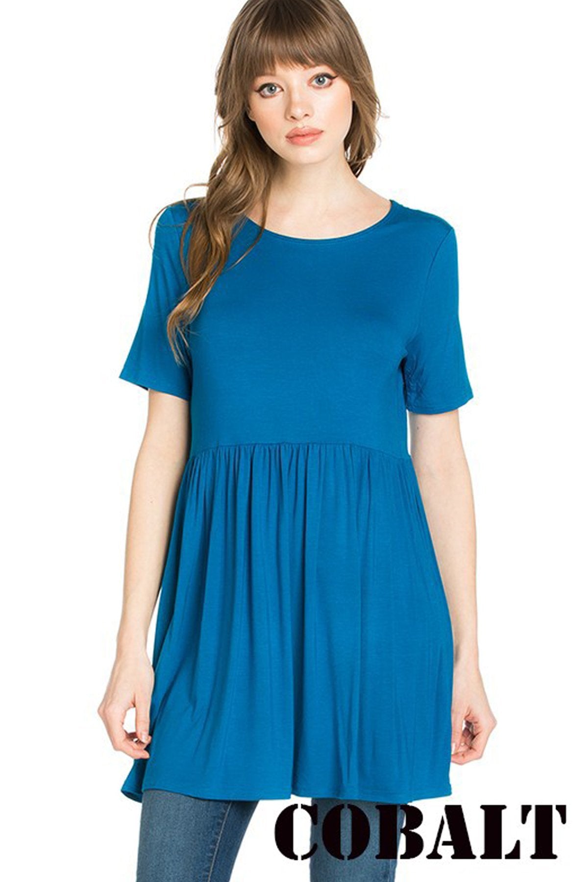 A woman stands outdoors wearing a beige Riah Fashion Short Sleeve Empire Waist Tunic paired with a long necklace.