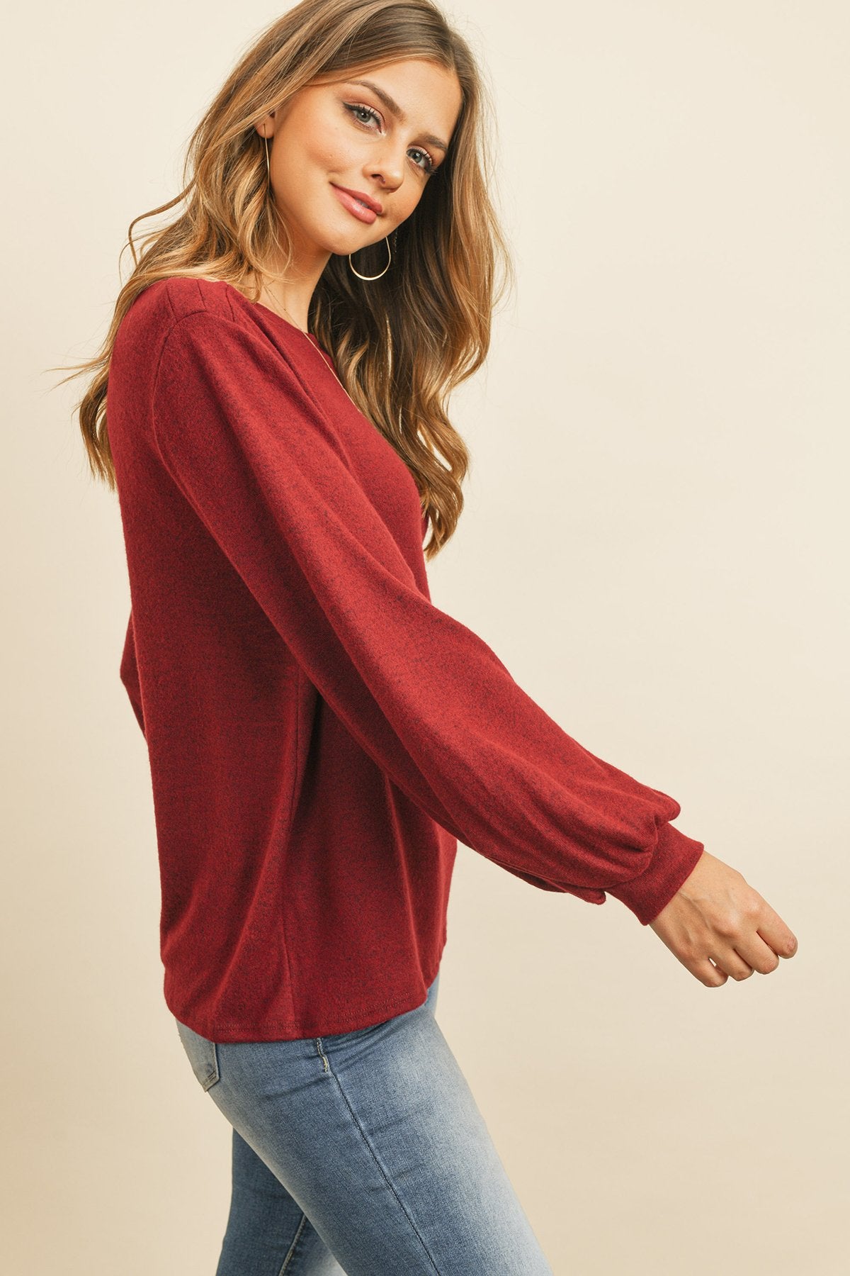 A woman models a Riah Fashion Puff Sleeved Boat Neck Two Toned Brushed Hacci Top with ripped jeans and brown boots on a concrete floor against a plain backdrop, accessorized with a long necklace.