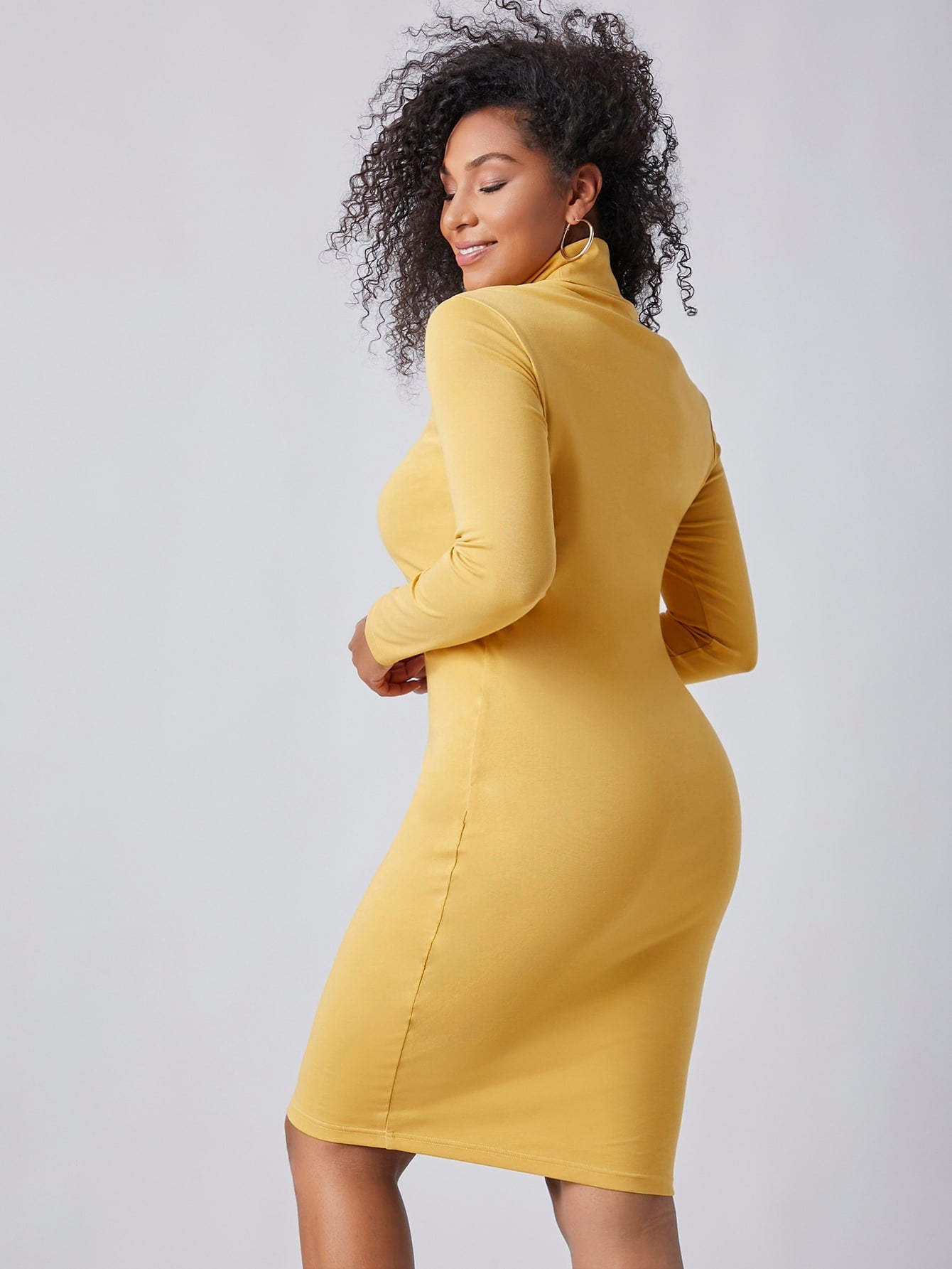 A person models wearing the Bright & Delight Spring Dress, a fitted yellow turtleneck dress from BCNY BOUTIQUE, posing with one hand on their hip and the other near their face against a plain background.