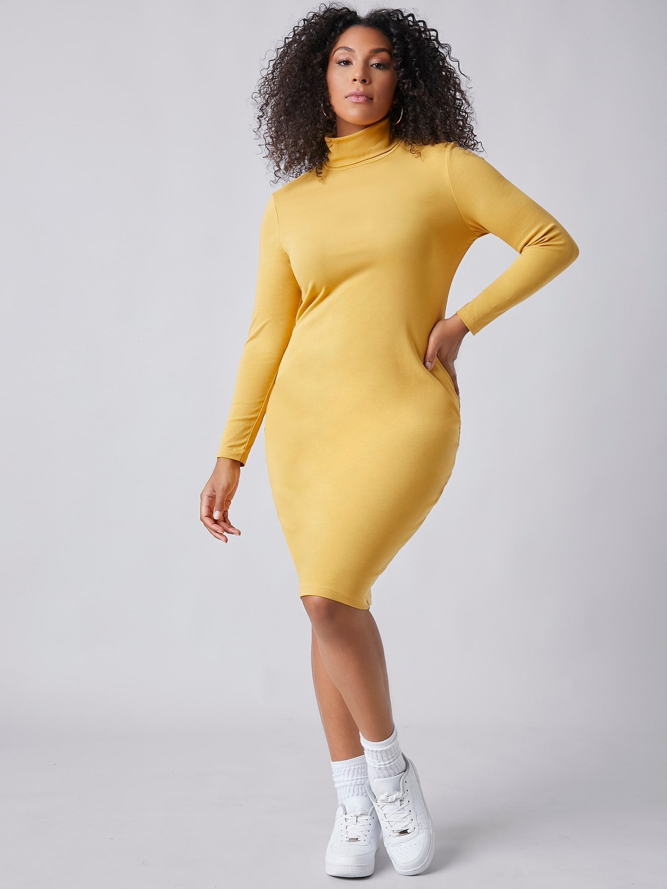 A person models wearing the Bright & Delight Spring Dress, a fitted yellow turtleneck dress from BCNY BOUTIQUE, posing with one hand on their hip and the other near their face against a plain background.