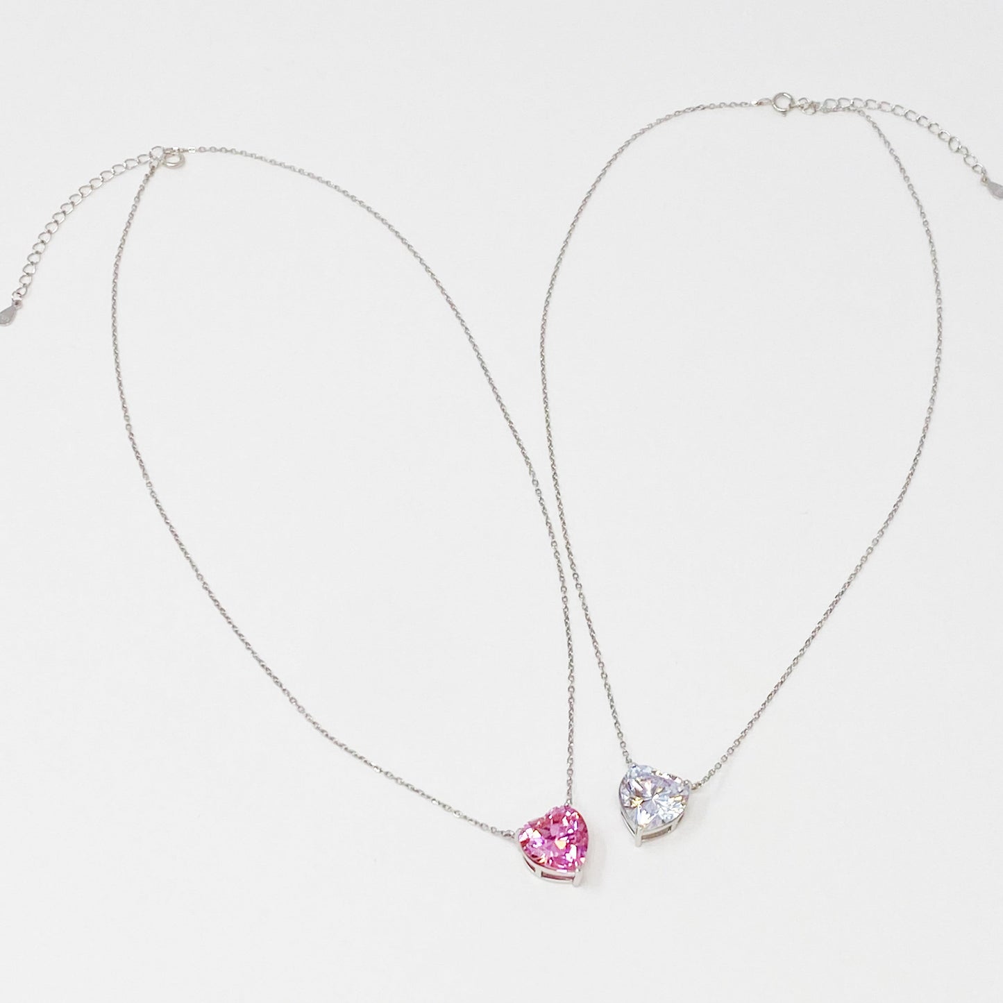 Two Absolute Beauty sterling silver heart necklaces by Ellison + Young; one with a pink gem, the other with a clear gem, against a white background.