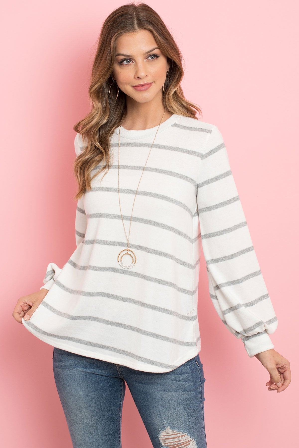A woman in a Riah Fashion Striped Long Sleeved Round Neck Swing Top and jeans stands against a pink backdrop, wearing a long necklace and touching her hair.