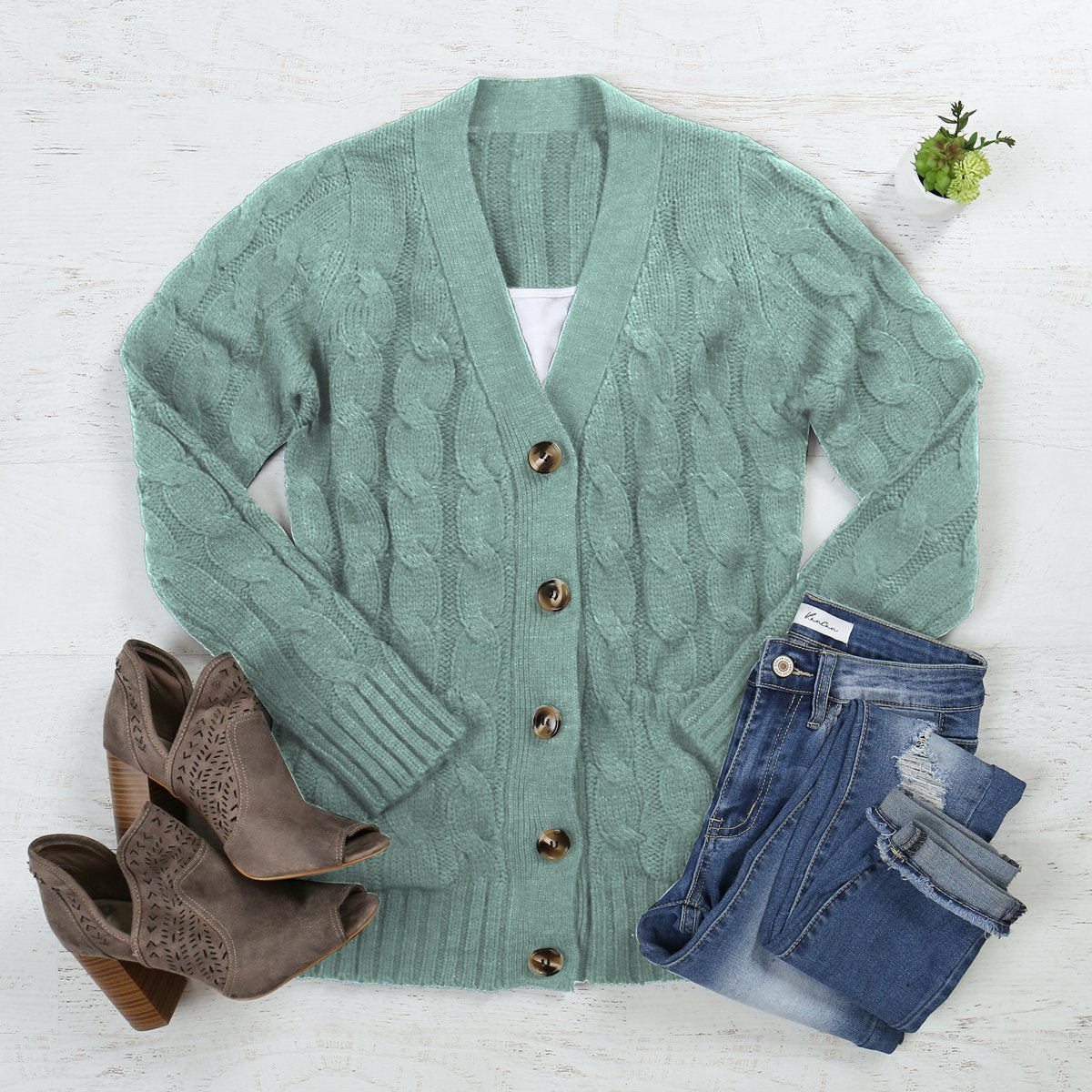 A person wearing an ash copper cable sweater with pockets from Riah Fashion stands in front of tall green plants and a wooden fence.