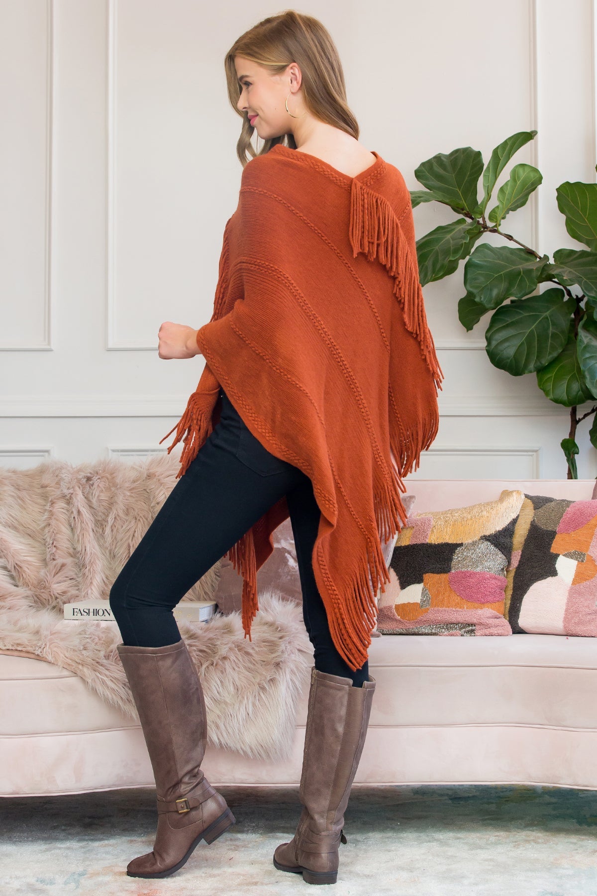 A person stands wearing a Riah Fashion Asymmetric Fringe Poncho in one color, paired with jeans and brown boots, smiling while holding the poncho. In the background are a plant and a sofa with cushions.