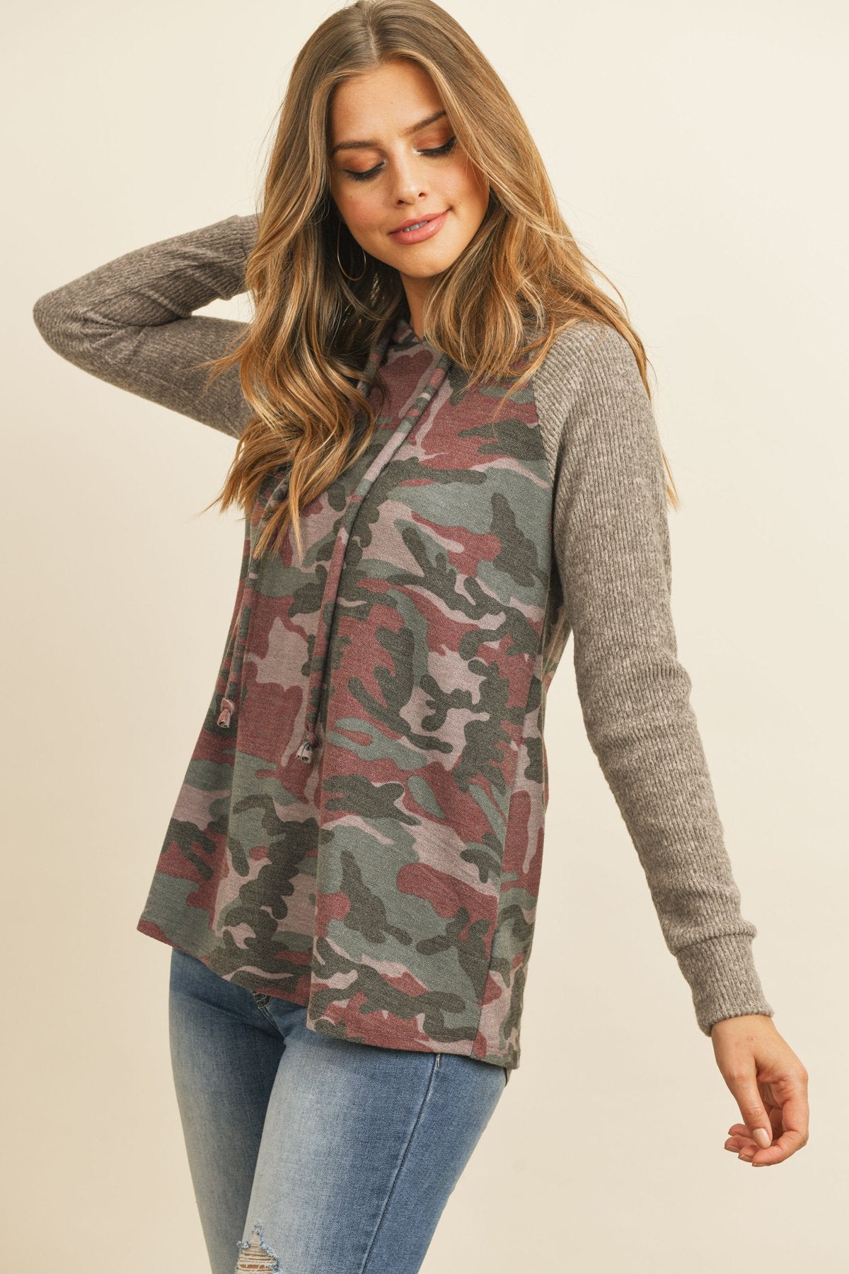 A person wearing the Self Tie Rib Hacci Brushed Sleeve Camouflage Hoodie by Riah Fashion with distressed jeans, holding the hoodie strings, stands against a beige background.