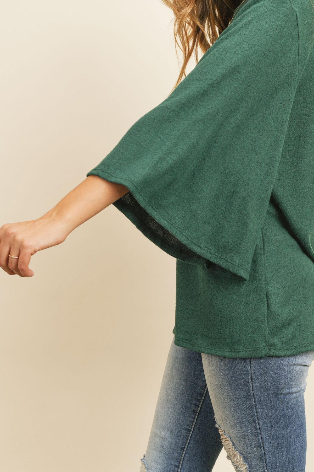 A person in a Riah Fashion Boat Neck Bell Sleeve Solid Hacci Brushed Top and ripped jeans stands against a neutral background, accessorized with hoop earrings and a long necklace.