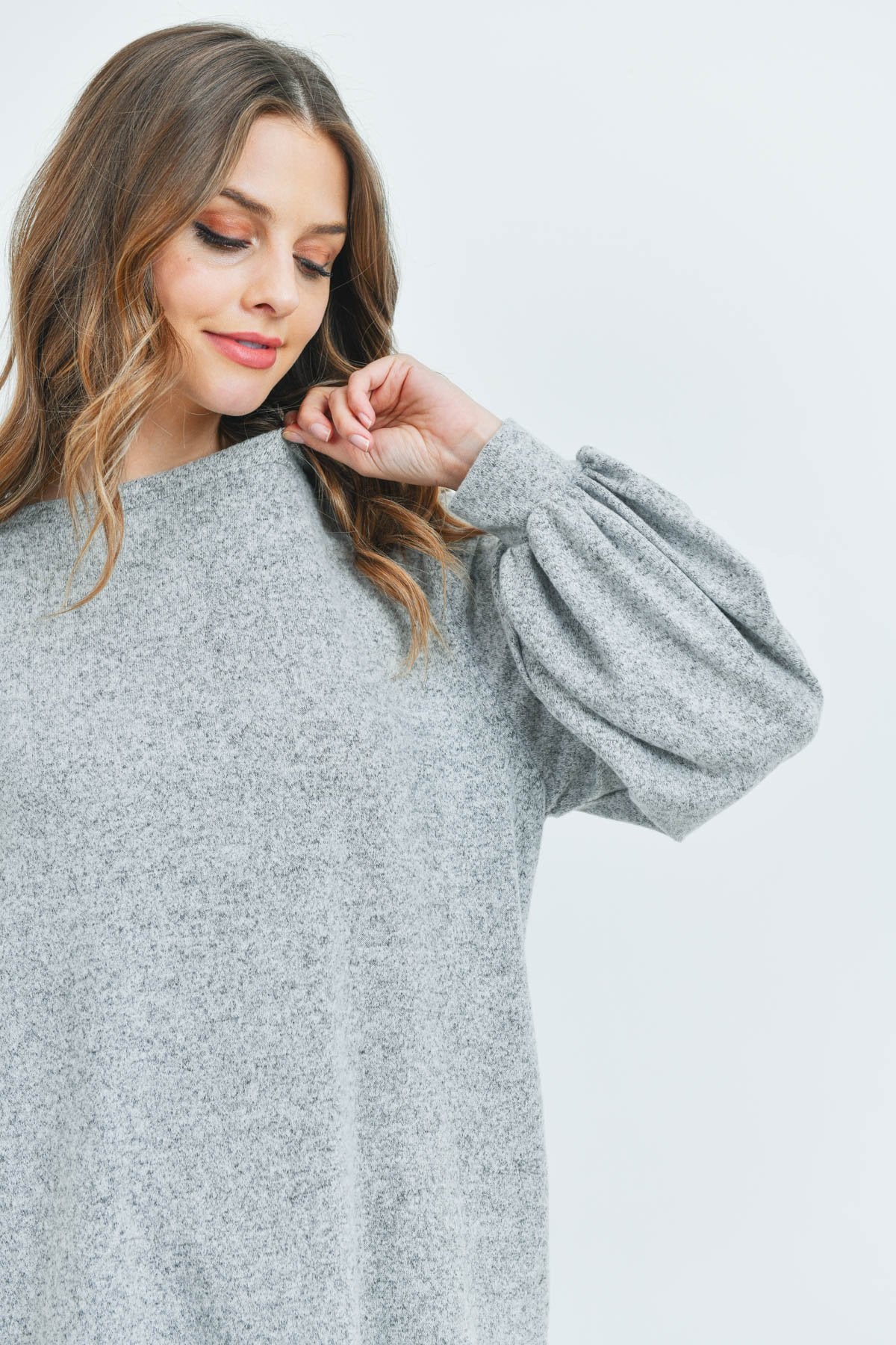 A woman models a Riah Fashion Puff Sleeved Boat Neck Two Toned Brushed Hacci Top with ripped jeans and brown boots on a concrete floor against a plain backdrop, accessorized with a long necklace.
