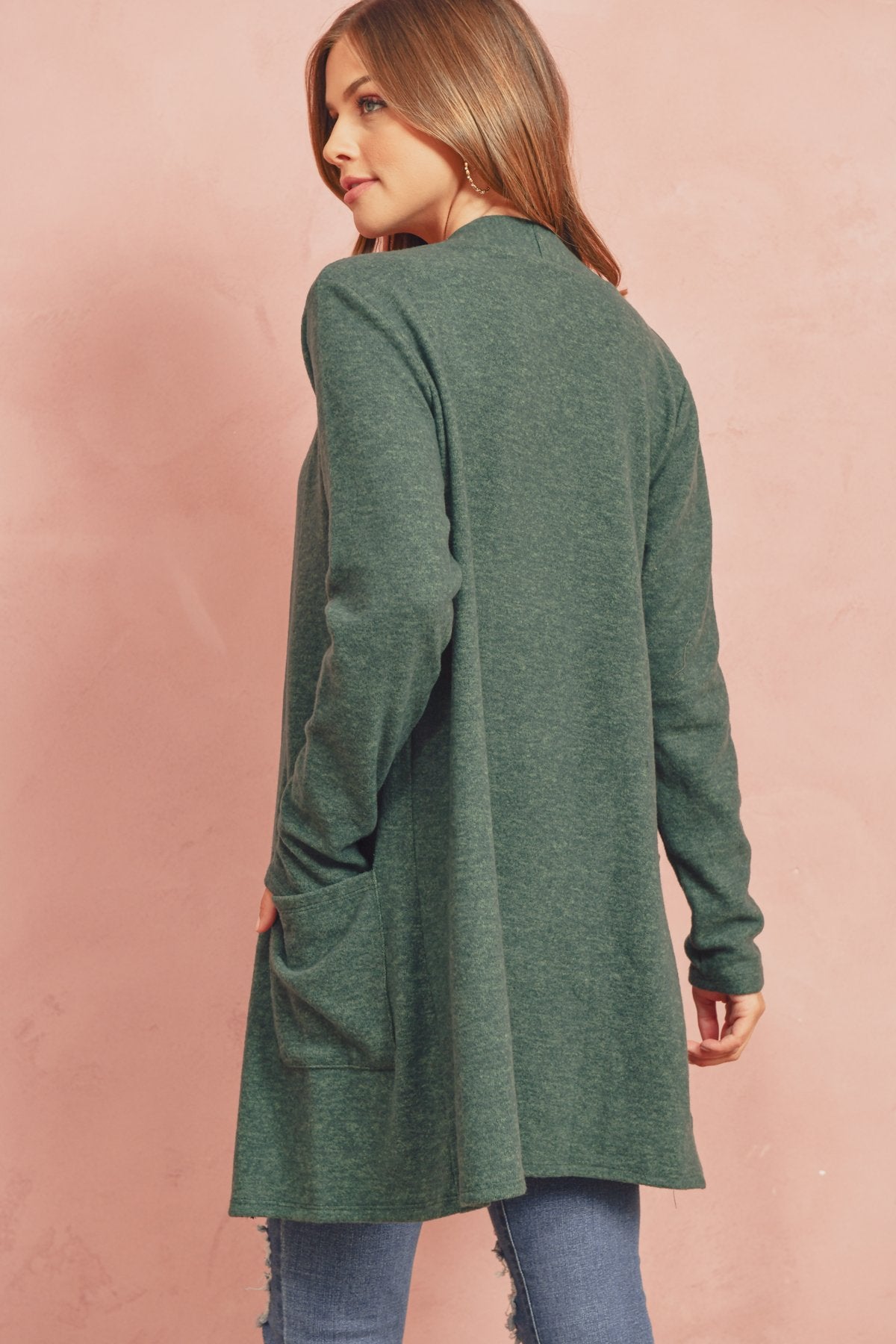 A woman in a Brushed Melange Open Front Cardigan by Riah Fashion and ripped jeans stands against a pink background, hands in her pockets.