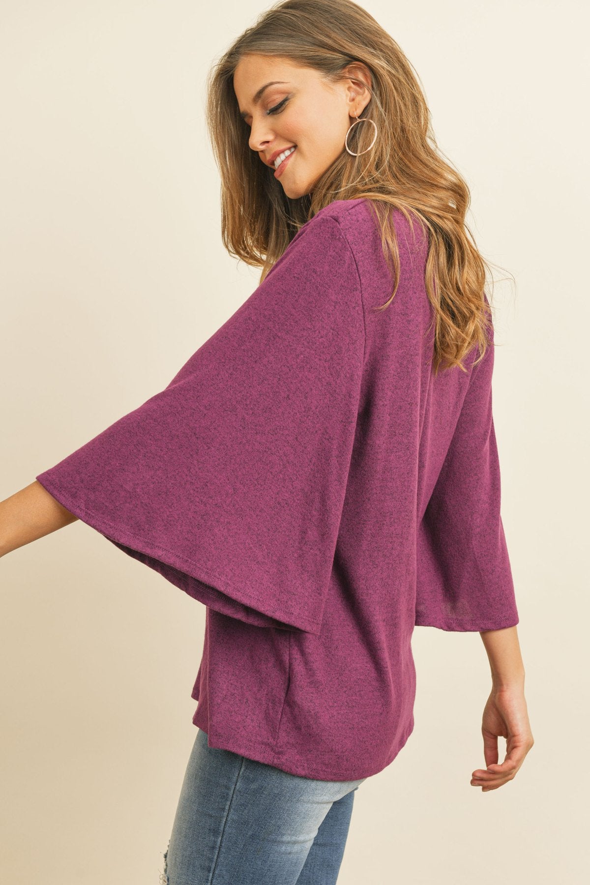 A person in a Riah Fashion Boat Neck Bell Sleeve Solid Hacci Brushed Top and ripped jeans stands against a neutral background, accessorized with hoop earrings and a long necklace.