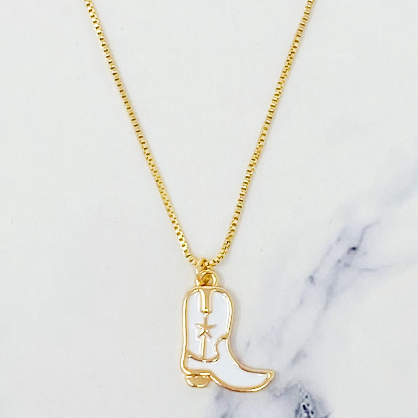 Two Cow Girl Boot Pendant Necklaces by Ellison + Young, one with a white boot and the other with a pink boot, are displayed on a marble surface.