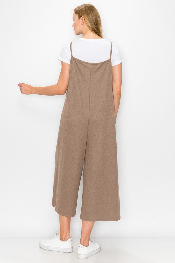 A person with long blonde hair sits on a wooden stool against a plain background, wearing Stylespects Cropped Bottom Wide Leg Oversized Jumpsuit in taupe and white sneakers.