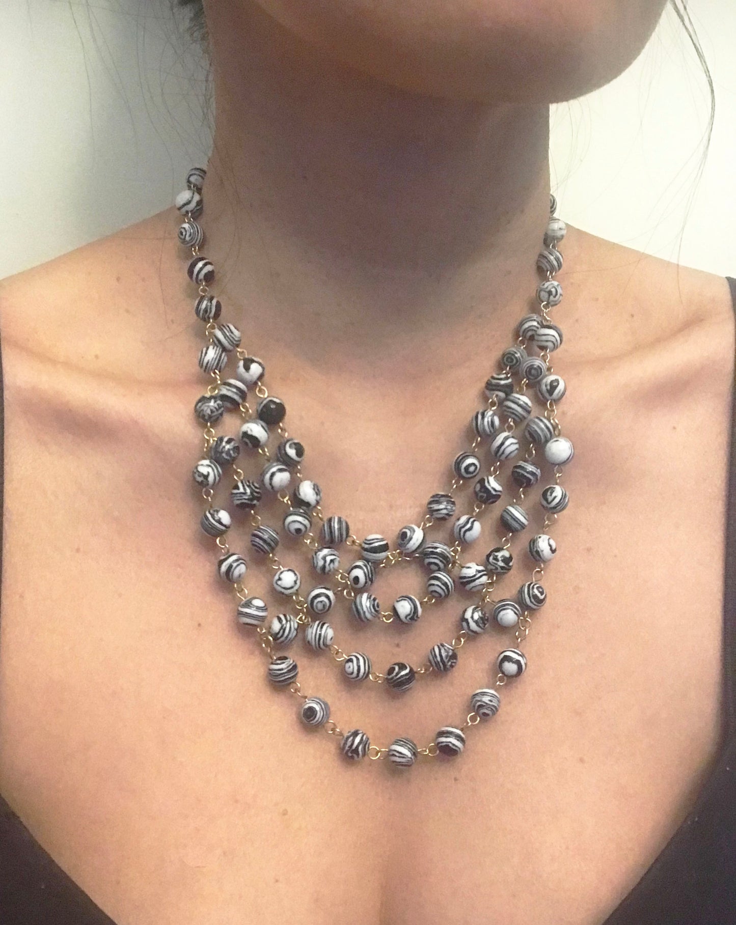 The Gena Myint Zebra Stripe Turquoise Statement Necklace features multi-strand black and white swirled beads with gold links, displayed on a white background.