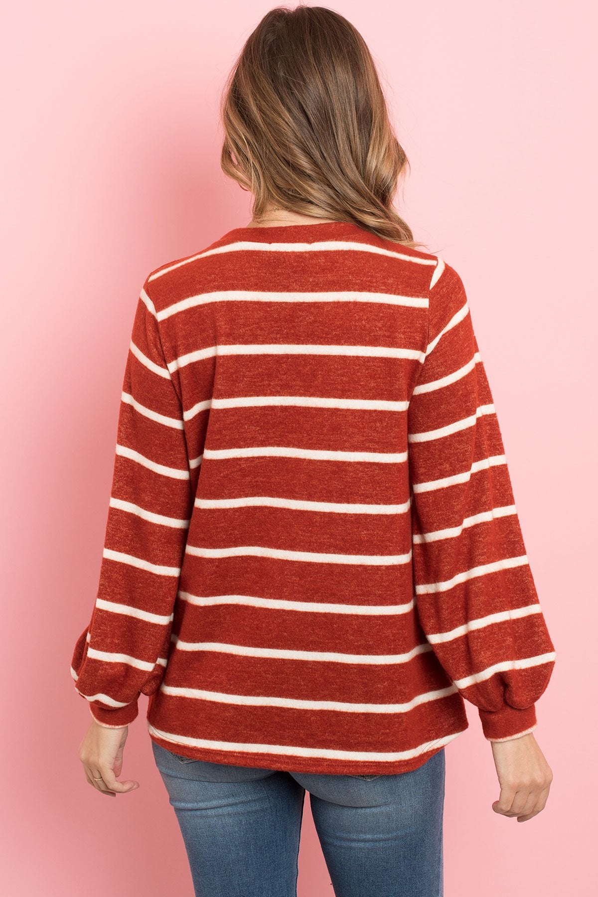 A woman in a Riah Fashion Striped Long Sleeved Round Neck Swing Top and jeans stands against a pink backdrop, wearing a long necklace and touching her hair.