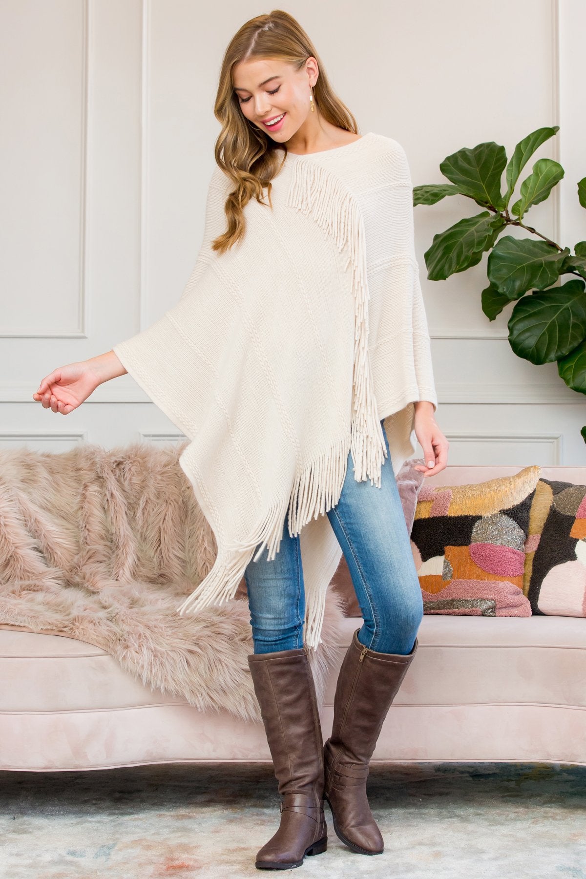 A person stands wearing a Riah Fashion Asymmetric Fringe Poncho in one color, paired with jeans and brown boots, smiling while holding the poncho. In the background are a plant and a sofa with cushions.