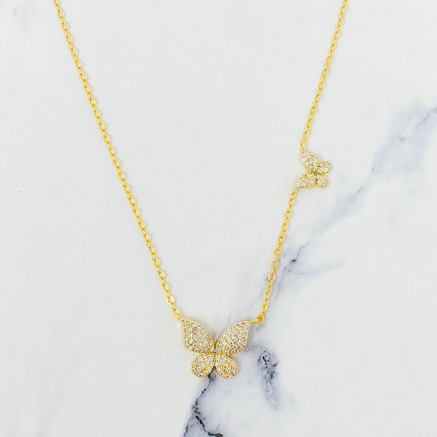 The Ellison + Young Double Shine Butterfly Necklace, featuring butterfly-shaped pendants adorned with small crystals, is elegantly displayed on a light marble background.