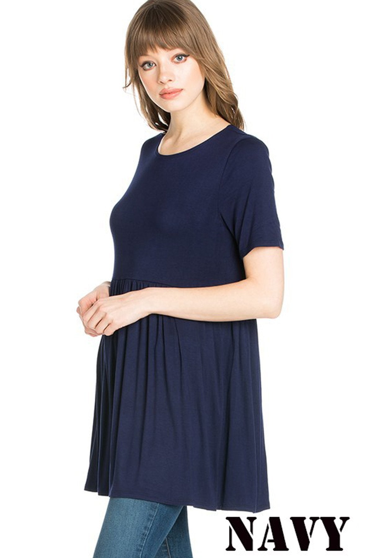 A woman stands outdoors wearing a beige Riah Fashion Short Sleeve Empire Waist Tunic paired with a long necklace.