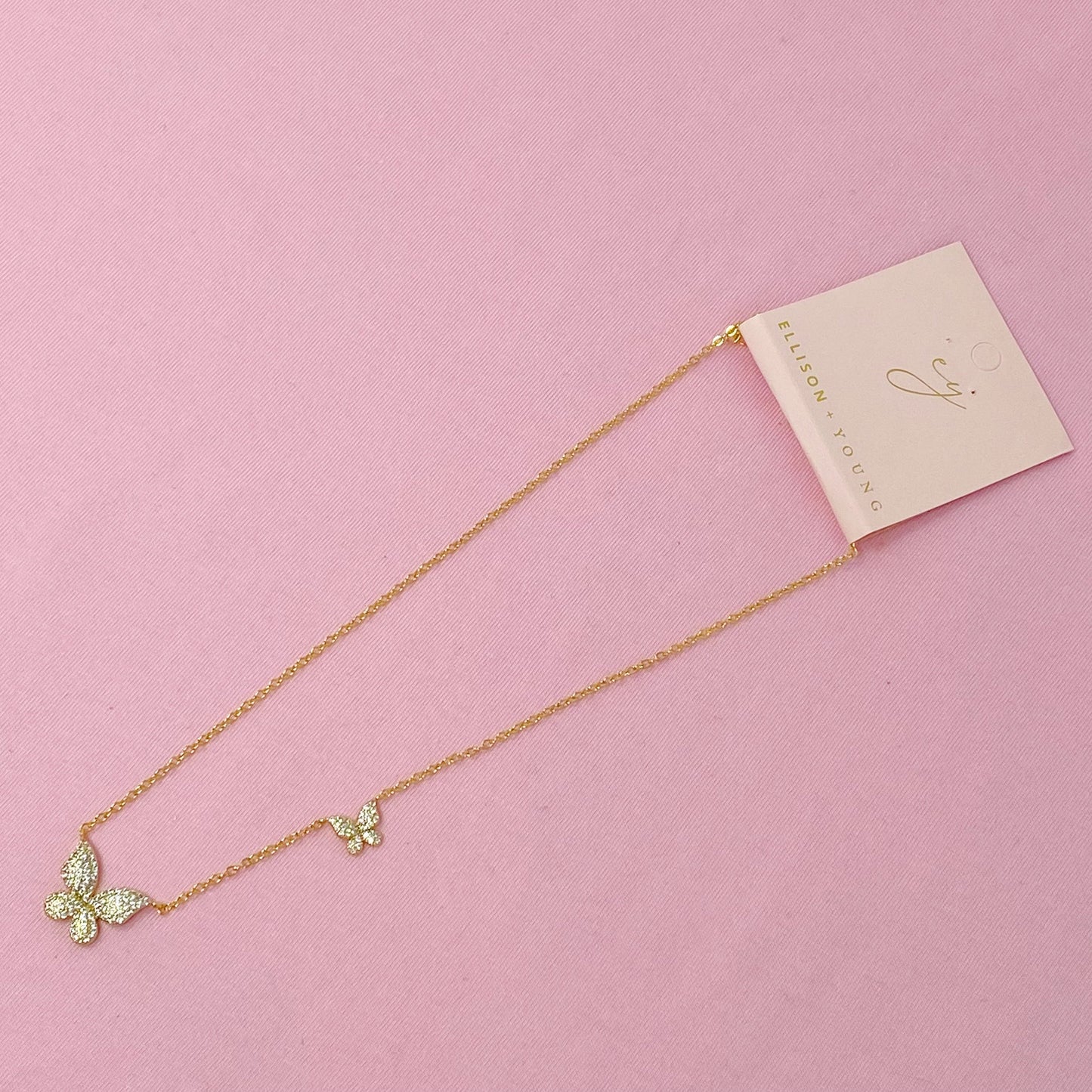 The Ellison + Young Double Shine Butterfly Necklace, featuring butterfly-shaped pendants adorned with small crystals, is elegantly displayed on a light marble background.
