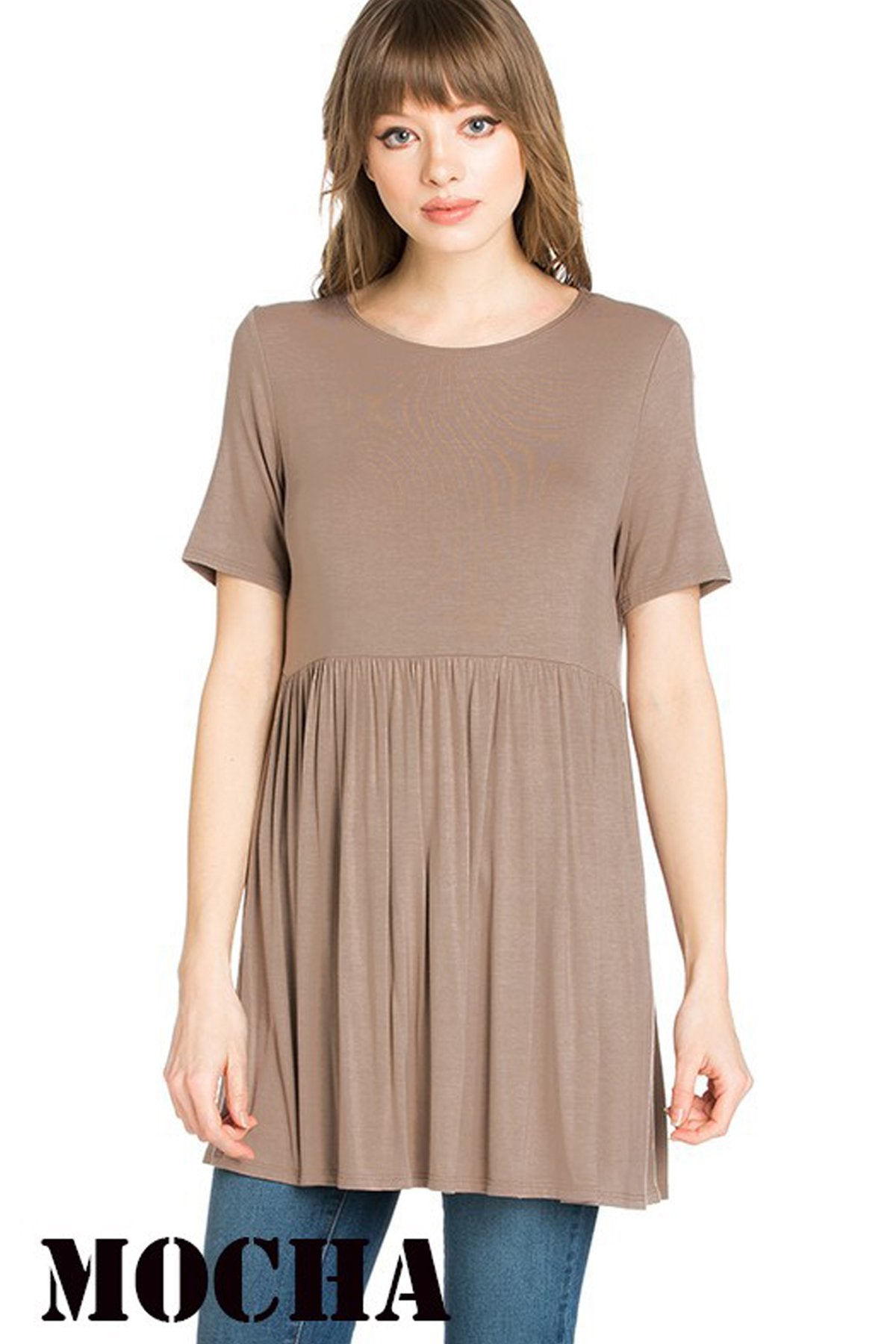 A woman stands outdoors wearing a beige Riah Fashion Short Sleeve Empire Waist Tunic paired with a long necklace.