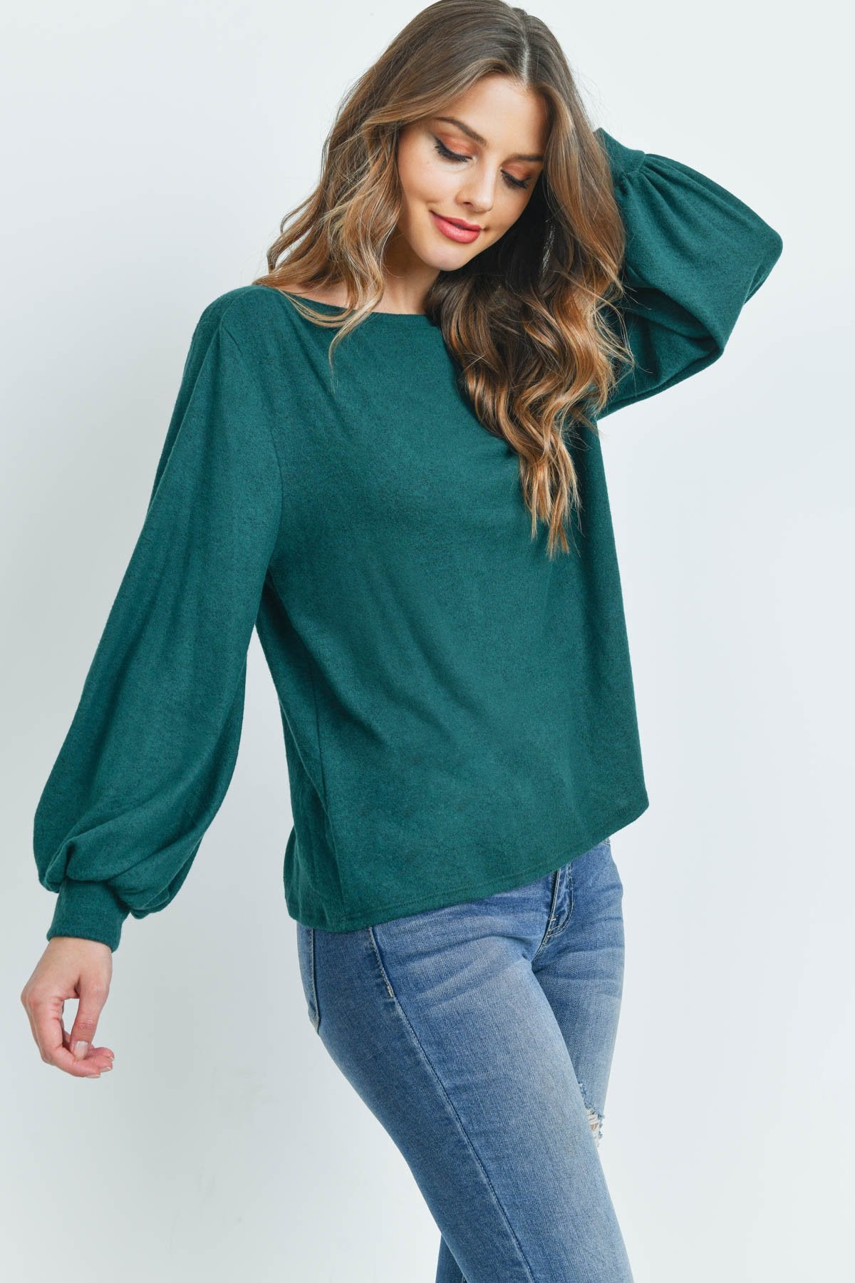 A woman models a Riah Fashion Puff Sleeved Boat Neck Two Toned Brushed Hacci Top with ripped jeans and brown boots on a concrete floor against a plain backdrop, accessorized with a long necklace.