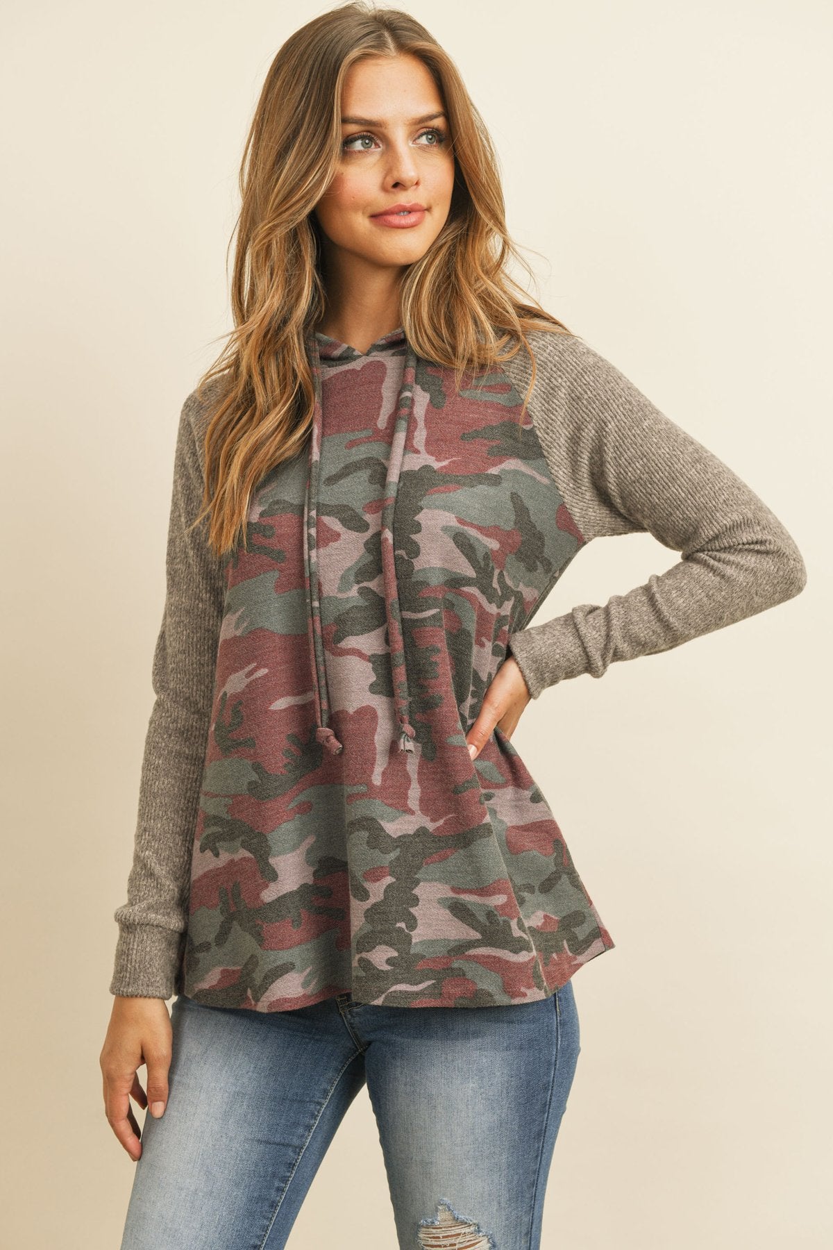 A person wearing the Self Tie Rib Hacci Brushed Sleeve Camouflage Hoodie by Riah Fashion with distressed jeans, holding the hoodie strings, stands against a beige background.