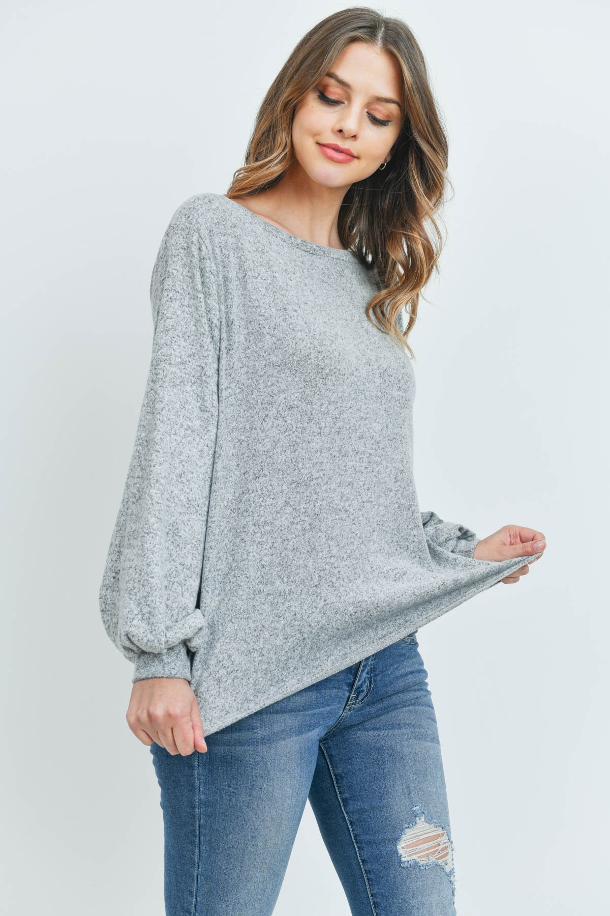 A woman models a Riah Fashion Puff Sleeved Boat Neck Two Toned Brushed Hacci Top with ripped jeans and brown boots on a concrete floor against a plain backdrop, accessorized with a long necklace.