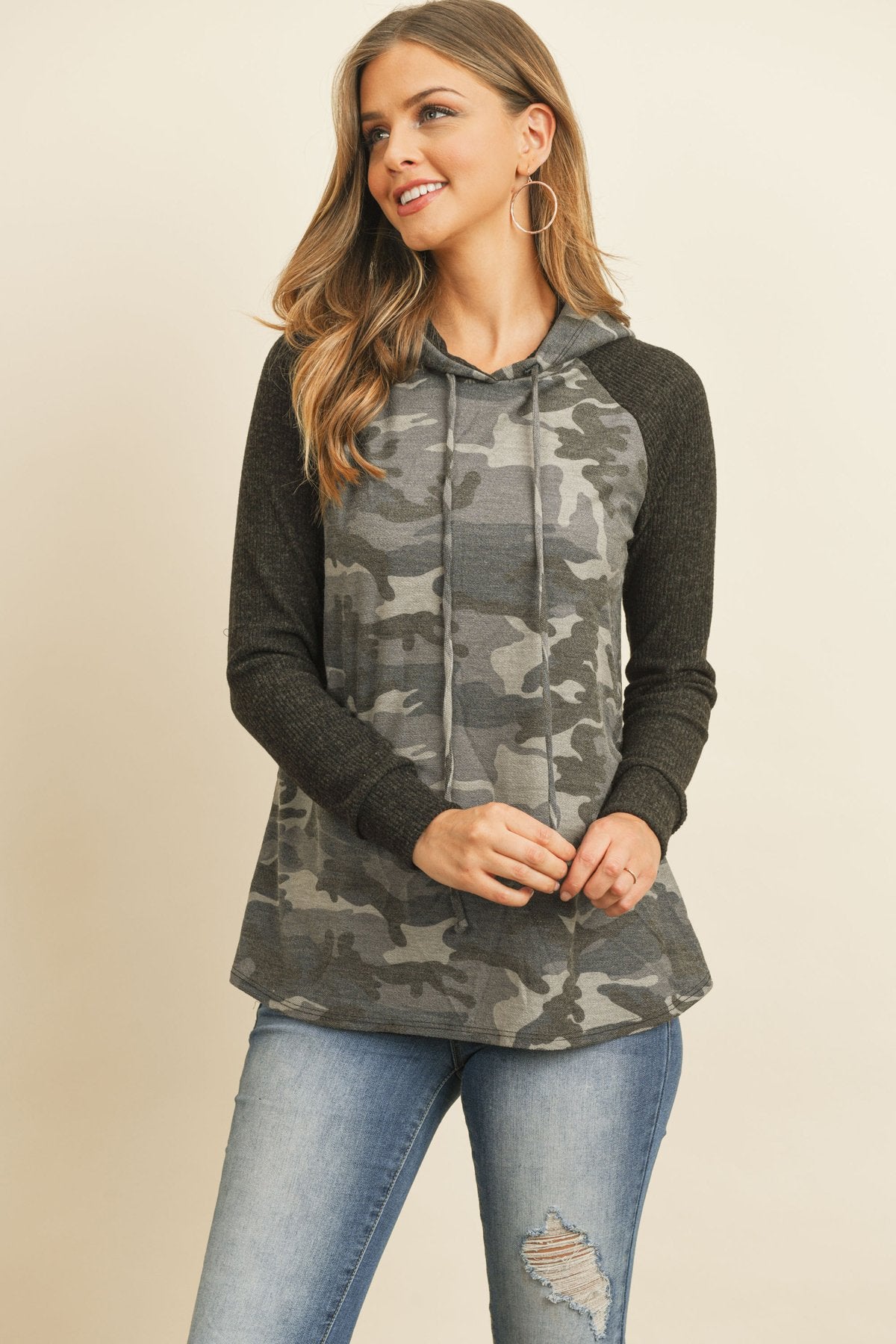 A person wearing the Self Tie Rib Hacci Brushed Sleeve Camouflage Hoodie by Riah Fashion with distressed jeans, holding the hoodie strings, stands against a beige background.