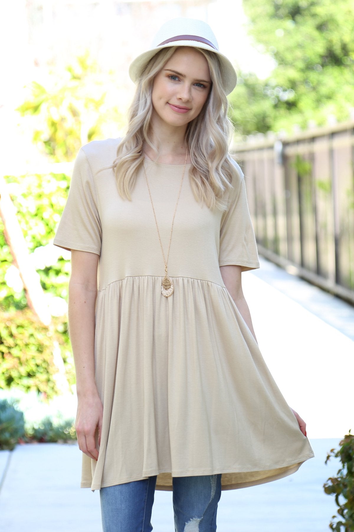 A woman stands outdoors wearing a beige Riah Fashion Short Sleeve Empire Waist Tunic paired with a long necklace.