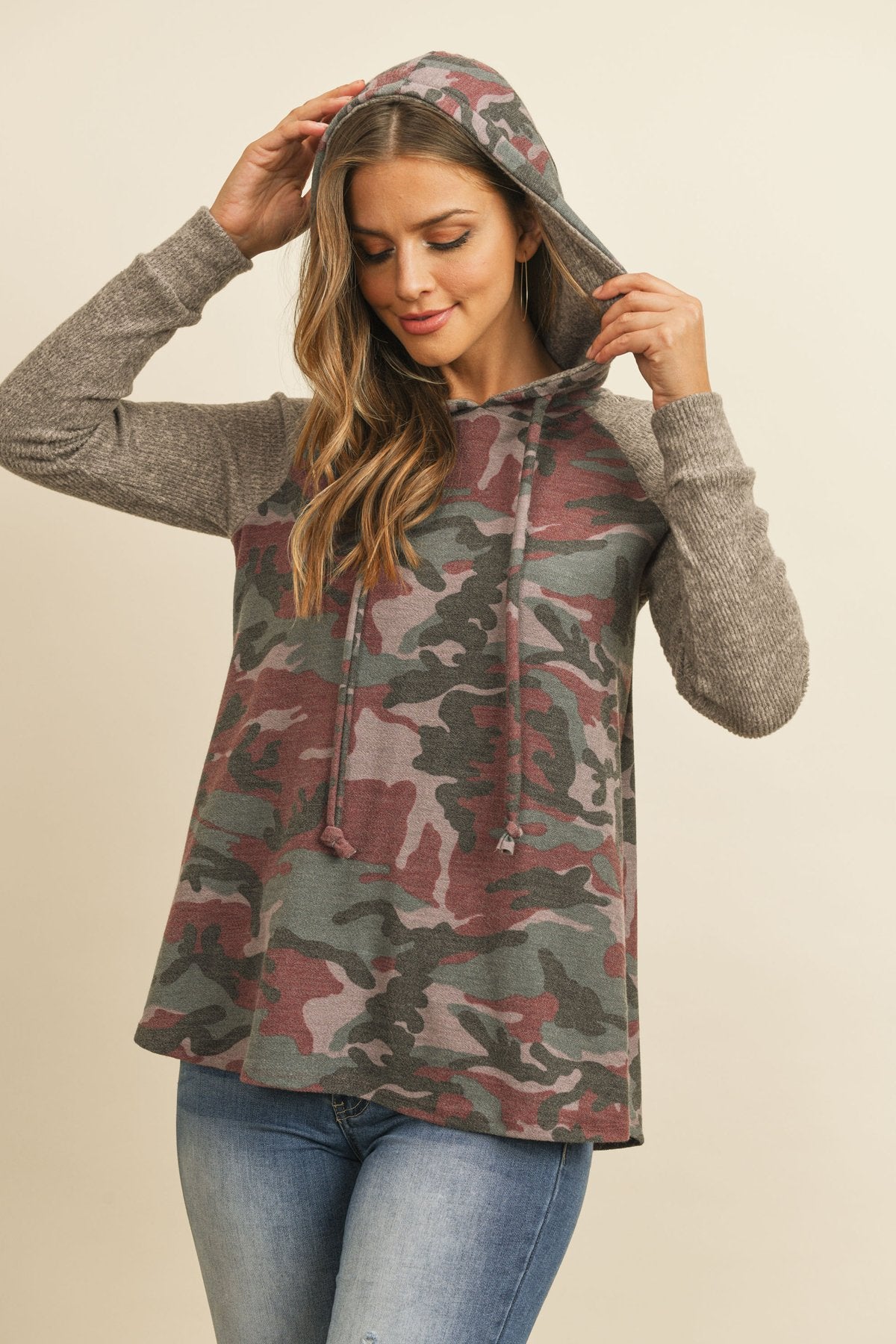 A person wearing the Self Tie Rib Hacci Brushed Sleeve Camouflage Hoodie by Riah Fashion with distressed jeans, holding the hoodie strings, stands against a beige background.