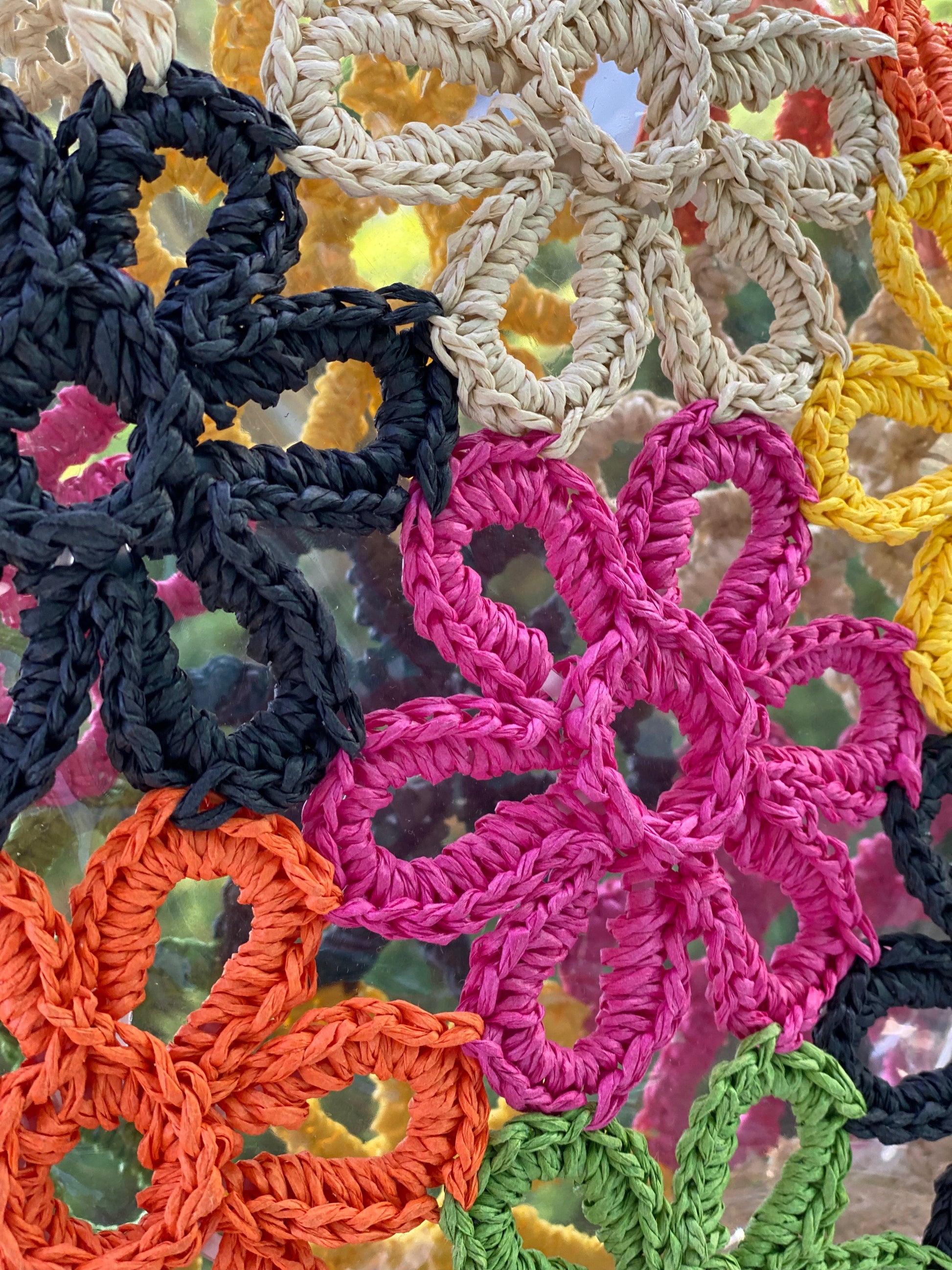 The Hand Made Flower Power Tote by Ellison + Young, featuring colorful floral crochet patterns in orange, green, pink, black, and yellow, rests on dense green foliage.
