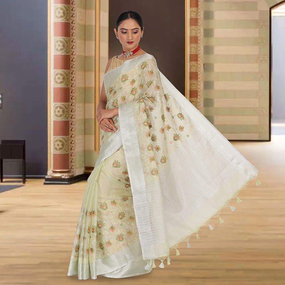 A woman in Chiros Tissue Banarasi Saree With Embroidery - Sea Green stands indoors on a wooden floor, against a decorative wall.