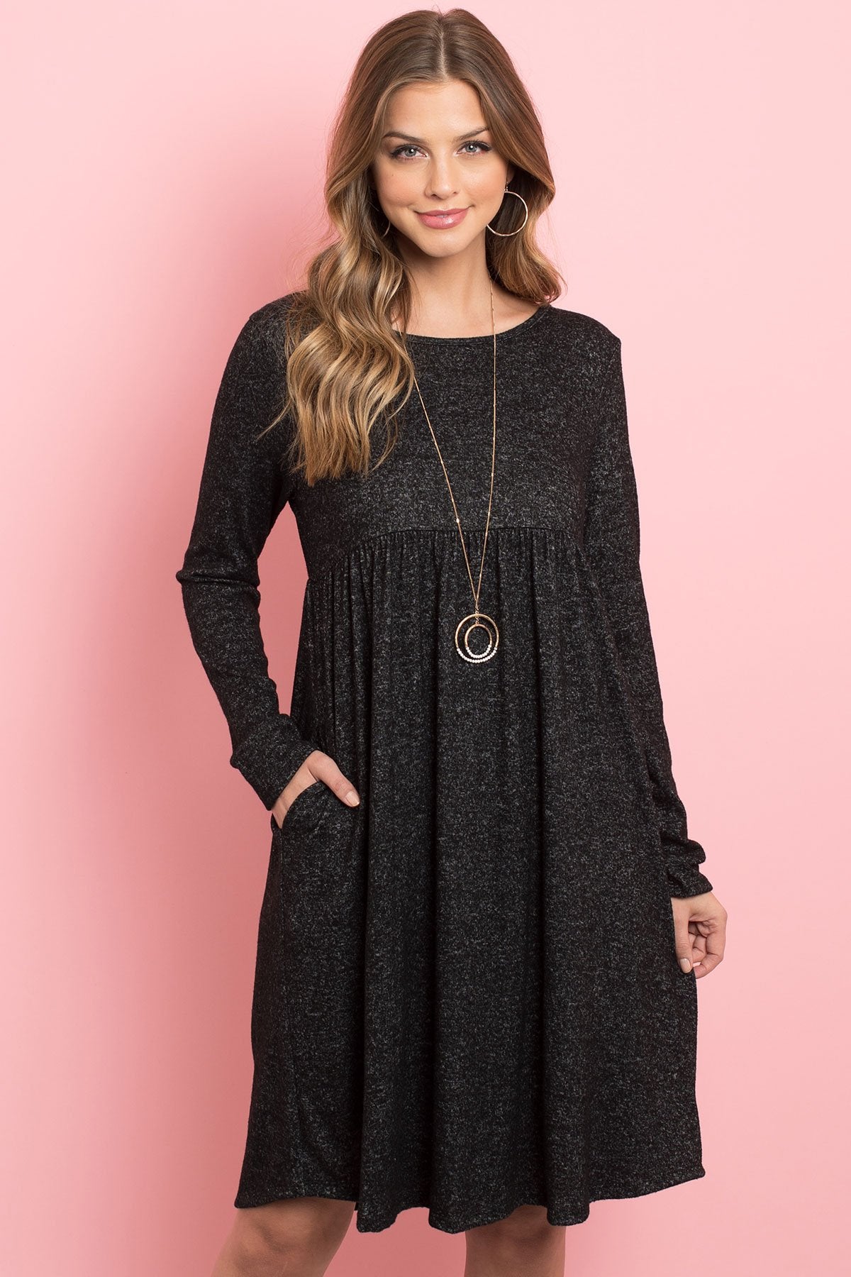 A woman in a Riah Fashion Boat Neck Brushed Hacci Round Hem Dress in Dark Ruby stands before a pink backdrop, adorned with a long necklace featuring circular pendants.