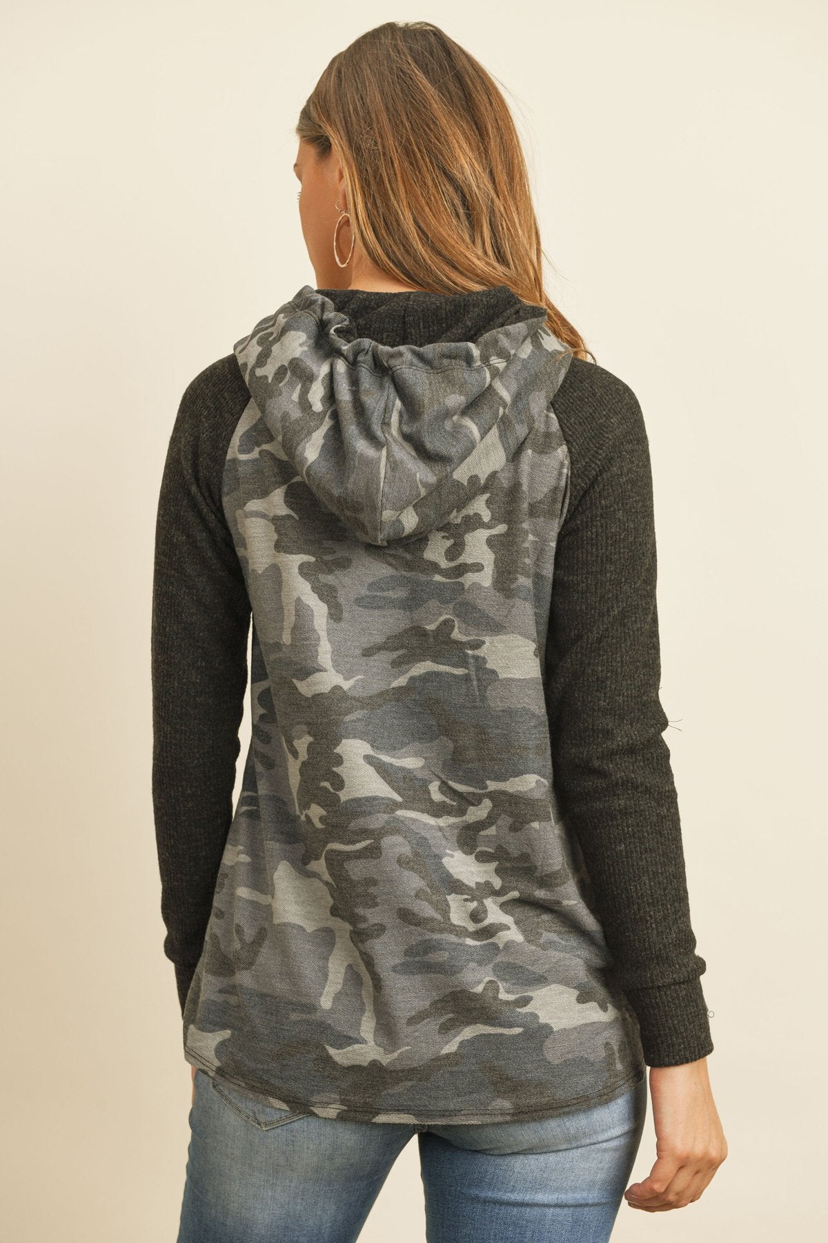 A person wearing the Self Tie Rib Hacci Brushed Sleeve Camouflage Hoodie by Riah Fashion with distressed jeans, holding the hoodie strings, stands against a beige background.