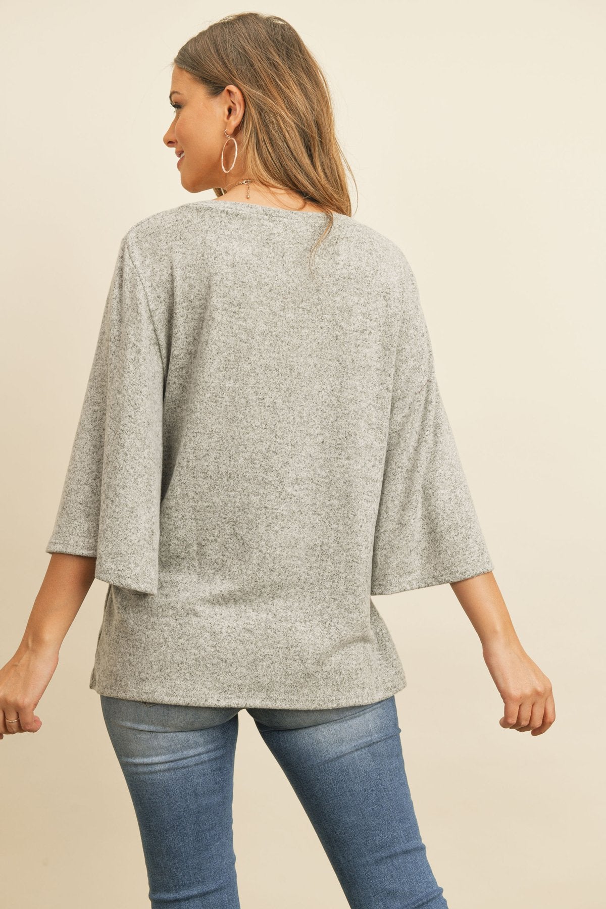 A person in a Riah Fashion Boat Neck Bell Sleeve Solid Hacci Brushed Top and ripped jeans stands against a neutral background, accessorized with hoop earrings and a long necklace.