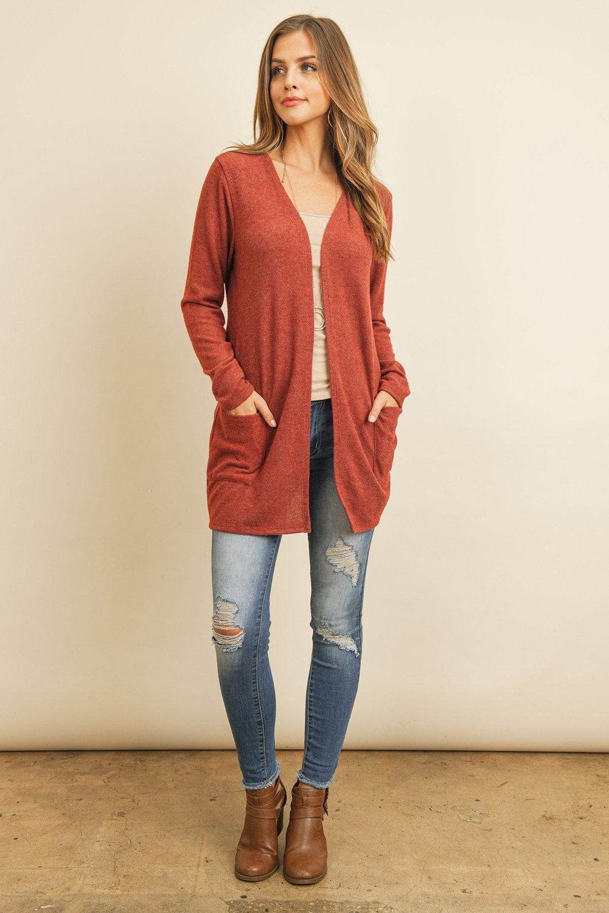 A woman in a teal Solid Long Sleeved Open Front Pocket Cardigan by Riah Fashion over a beige top stands against a pink background, smiling with hands in pockets.